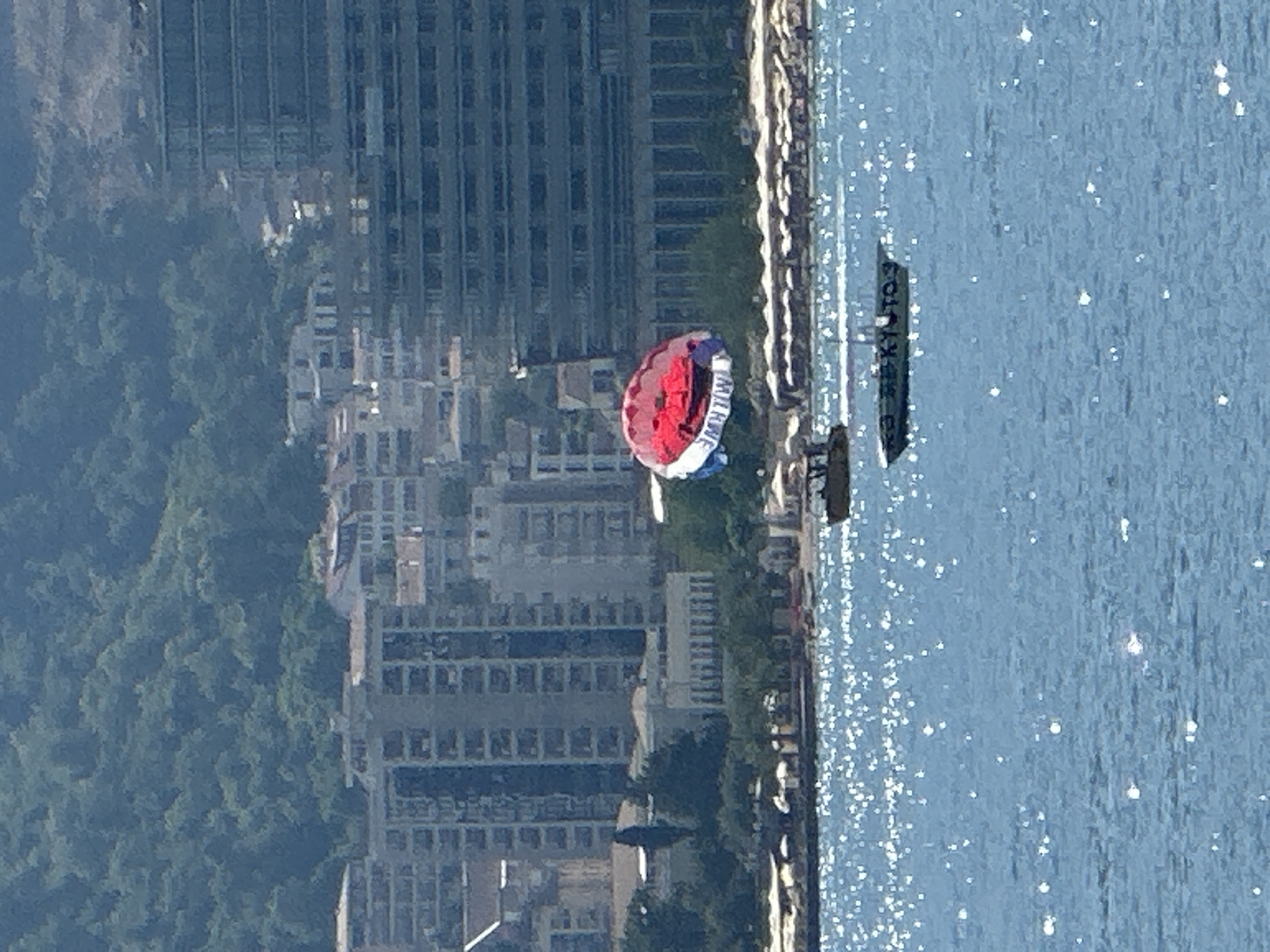 An aerial view captures a parasailer in mid-air above the Adriatic Sea, with a boat towing the parasailer visible in the water below. The parasail is a vibrant red, white, and blue. The coastline is lined with buildings, including high-rise apartments and hotels, with a lush, green hillside forming the backdrop. The sea is a bright blue, sparkling with sunlight. In the foreground, two boats are present on the water. One is a smaller, darker boat, likely the one towing the parasailer. The second is a longer, flat boat with text visible on its side. The scene appears to be a popular tourist area in Budva, Montenegro, indicated by the resort-style buildings and recreational water activities.