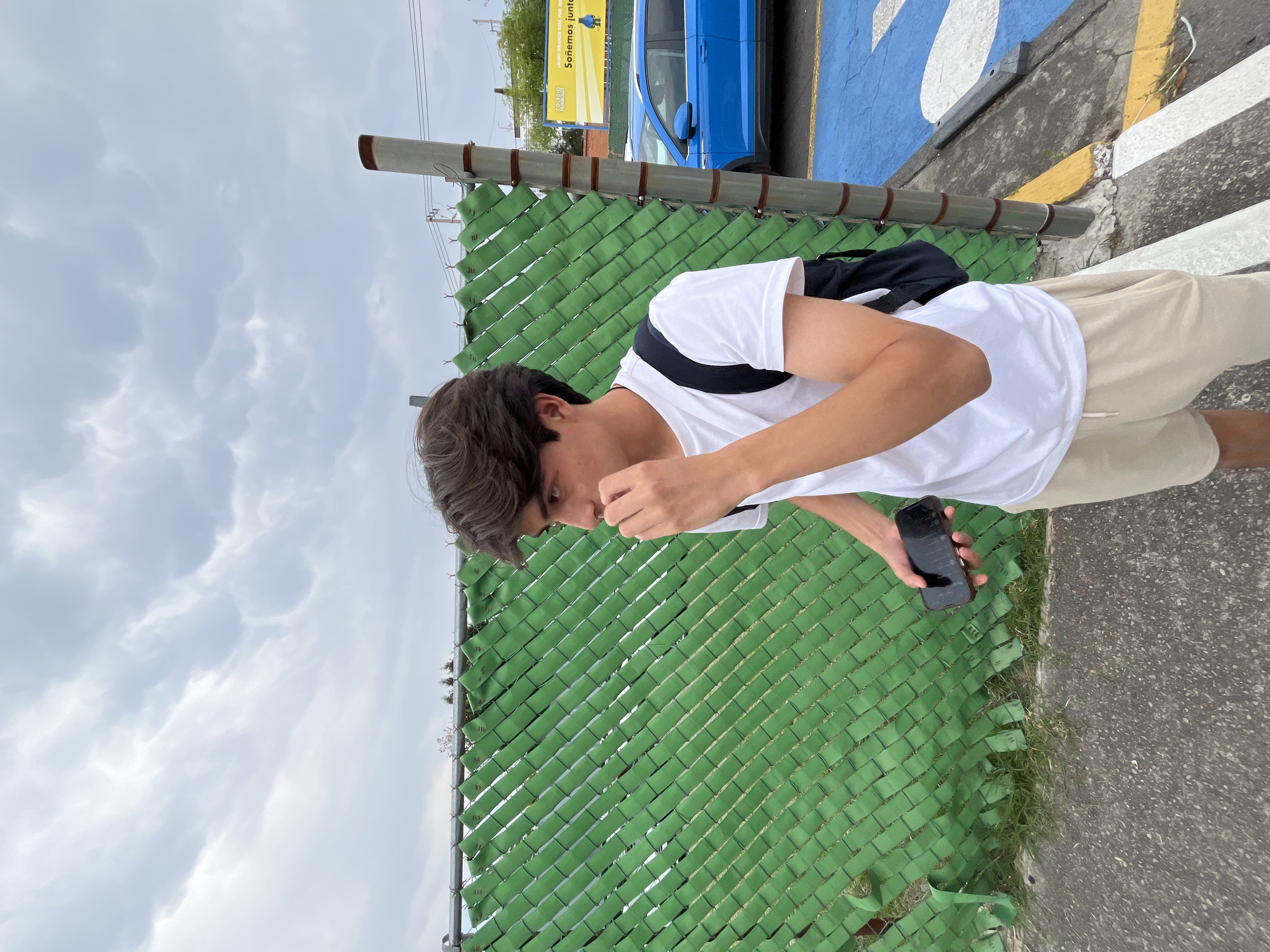A young man stands outdoors on a cloudy day in Puebla, Mexico, looking downwards at a mobile phone he holds in his left hand. He is wearing a white t-shirt, light-colored shorts, and a dark backpack, with his right hand raised near his face.
To his right, a tall green chain-link fence, fitted with privacy slats, runs horizontally across the midground. Beyond the fence, a paved area with distinct markings is visible, featuring a large blue section and white and yellow lines. A bright blue car is parked in the background, partially obscured by the fence and markings.
In the upper right background, a yellow billboard displays text. The visible Spanish phrases on the billboard are "EL VALOR GRANDE ESTÁ POR NACER" (The great value is yet to be born) and "Soñemos juntos" (Let's dream together), alongside a logo that includes the word "colegio" (school). The upper left portion of the image shows a sky filled with light grey clouds. The immediate ground surface where the man stands is asphalt, with some sparse grass growing along the base of the fence. The setting suggests an urban or suburban environment, potentially near a school or parking lot.