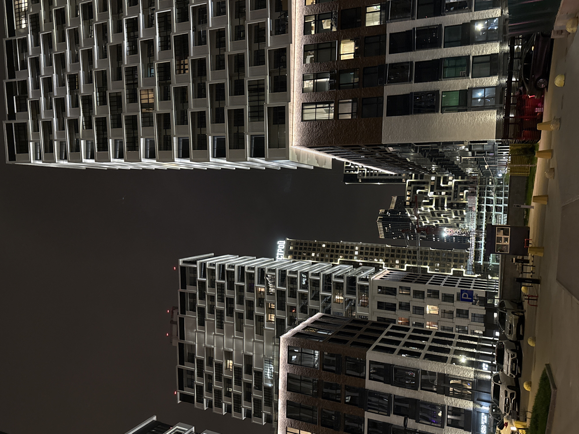 The image depicts a nighttime scene in Khimki, Russia, featuring several modern high-rise buildings. The buildings have distinctive architectural designs, including projecting window frames that create a textured facade. The buildings are lit from within, and the city lights reflect off the exterior. Some buildings have a white and grey color scheme, while others incorporate brown elements. A few cars are parked along a street beside one of the buildings, and there are some small barriers along the street. In the background, other buildings and construction cranes can be seen. The sky is dark, and the overall lighting is ambient from the city lights.