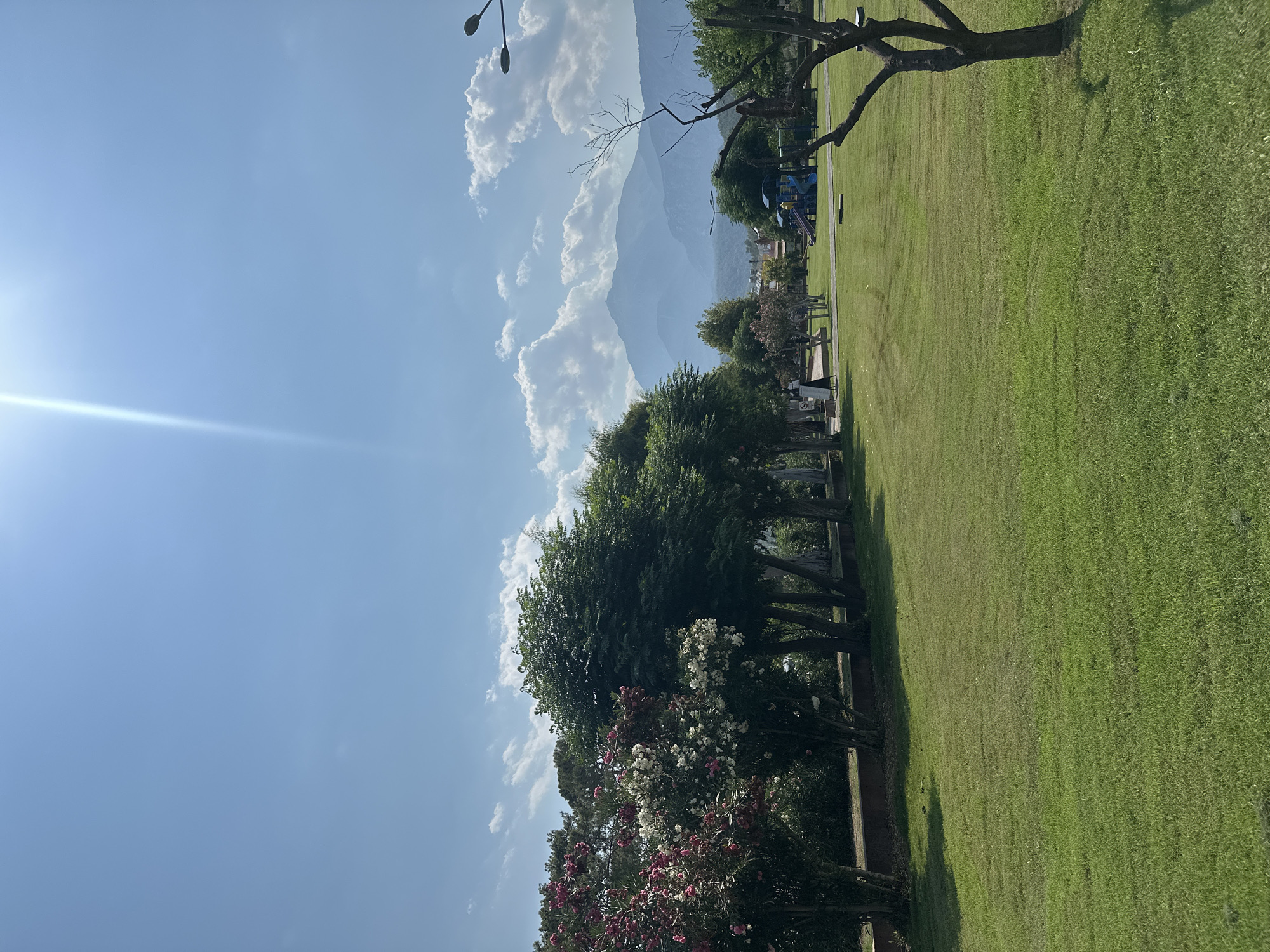This image depicts a park scene in Kemer, Turkey, under a clear blue sky with scattered white clouds. The foreground is dominated by a meticulously maintained green lawn. To the left, a line of trees with pink and white flowers forms a vibrant border, transitioning to taller, leafy green trees. In the middle ground, a playground with blue and yellow equipment is visible, with a few people walking in the distance. Beyond the playground, more trees and buildings are scattered. The background features a range of imposing mountains, shrouded in a light haze, indicating their distance. The overall impression is one of a peaceful, well-kept public space on a sunny day, with the natural beauty of the mountains providing a dramatic backdrop.