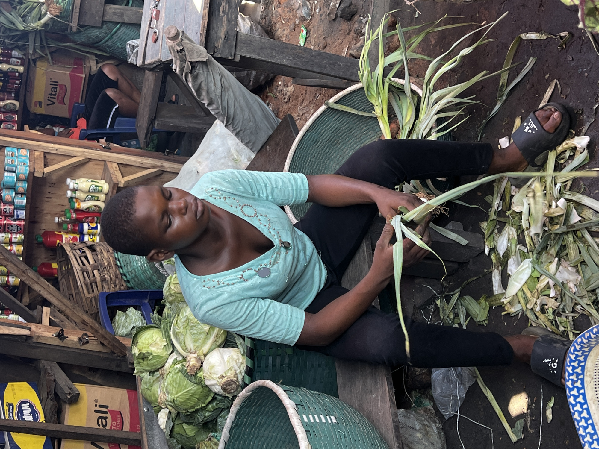 The image captures a vibrant, high-angle view of a woman working at a market stall in Nkpor, Nigeria, during the daytime. The main subject is a dark-skinned woman, seated on a low wooden structure, dressed in a light mint-green collared shirt with beaded embellishments and dark trousers, paired with black slides. She is actively engaged in preparing vegetables, holding several stalks of what appear to be leeks or similar leafy greens, with discarded outer layers scattered on the dirt ground around her. Her expression is focused as she works.
To her left, a pile of fresh cabbages, varying from light green to white, is visible next to a green woven basket. Another similar basket is positioned in front of her. The ground is bare earth, showing signs of wear and vegetable debris.
The background reveals the rustic construction of the market stall, made from rough wooden planks and beams. Shelves are stocked with various goods: rows of canned items (some blue, some red), bottles of condiments like ketchup and mayonnaise. Visible text includes "Vitali" on a ketchup box and "WHIP" on a mayonnaise box. A person wearing dark shorts is partially visible in the background, seated on a blue plastic chair, amidst boxes and other market items. The black slide on the woman's right foot has partially visible white text, likely "JEEP". The overall scene depicts a busy, open-air marketplace setting.