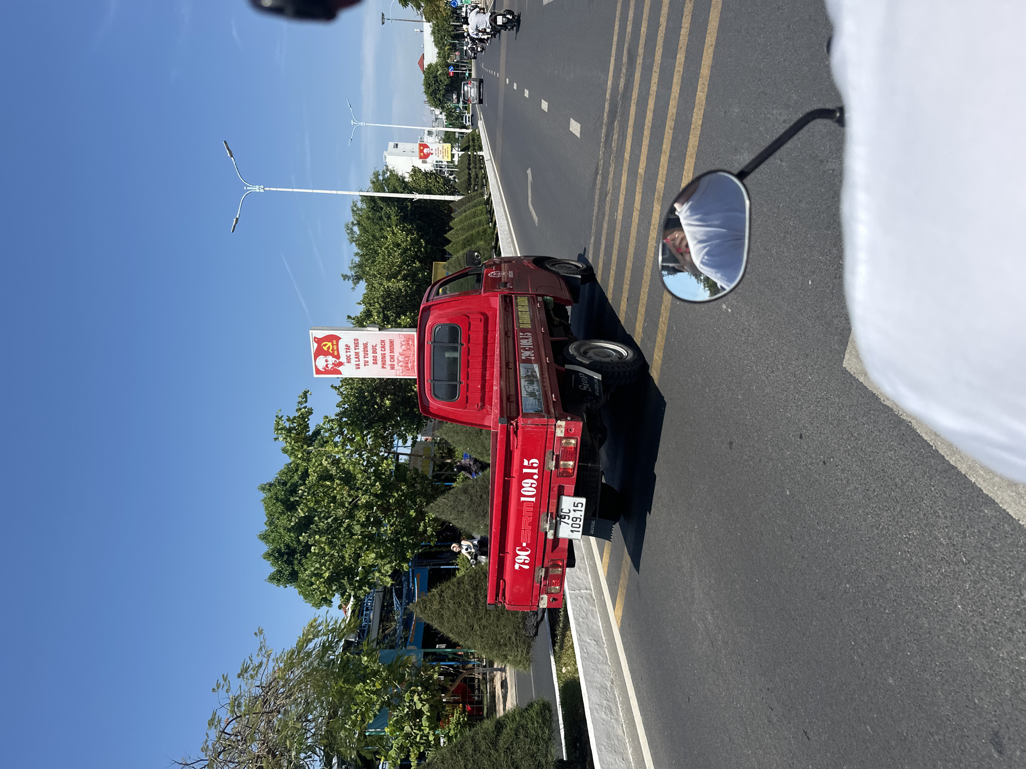 The image shows a red mini truck with the license plate "79C-SRM109.15" parked along the roadside in what appears to be Bac Nha Trang Ward, Vietnam. The truck is parked near a propaganda sign promoting study and following the ideology, ethics, and style of Ho Chi Minh. The sky is clear and blue. The image was likely taken from a passing motorcycle, as a motorcycle mirror and the rider's arm are visible in the shot. In the distance, there are more vehicles and buildings. Trees and bushes line the roadside. The focus is on the truck and its license plate.