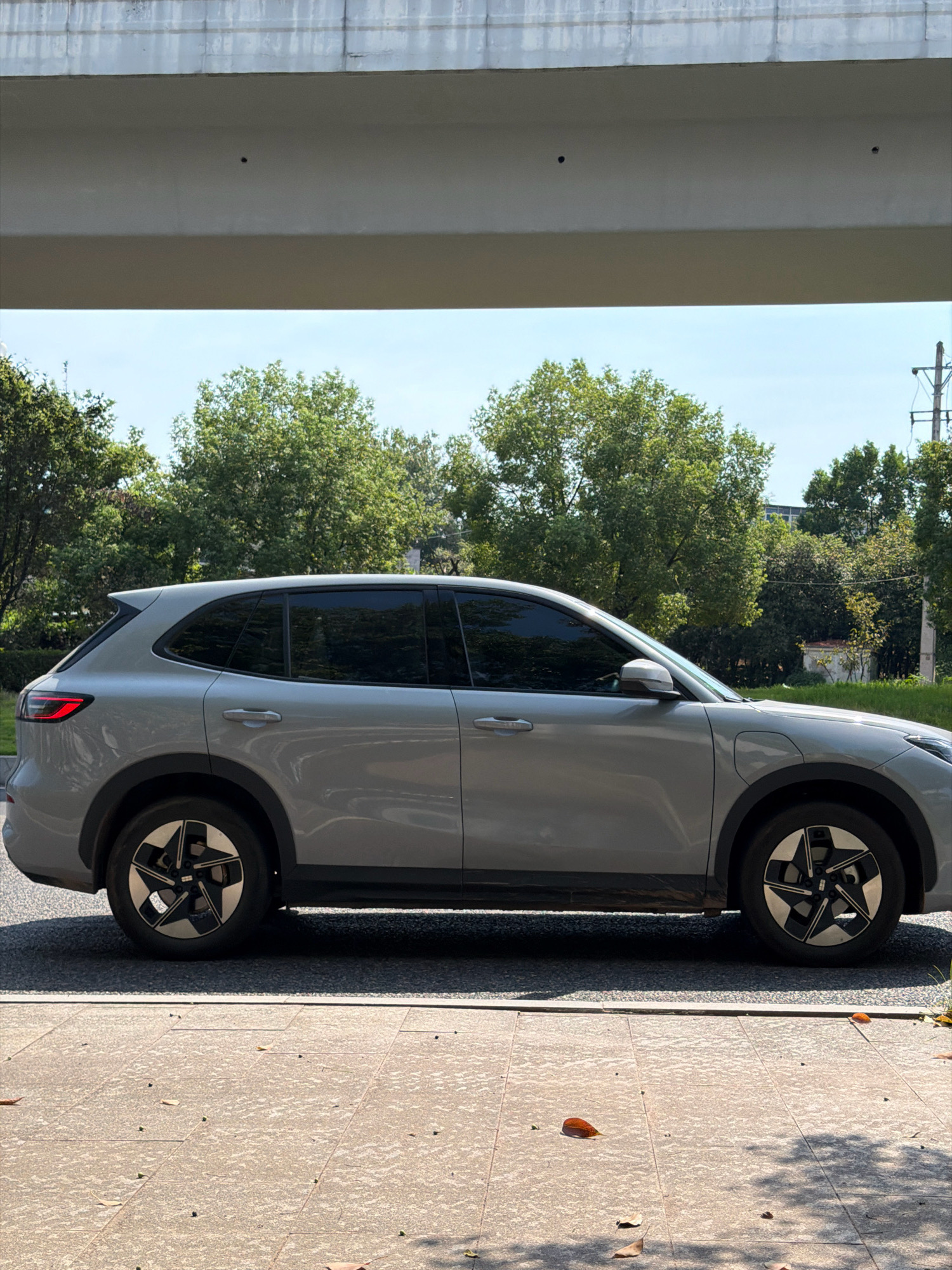 A silver electric SUV is parked on a paved road beneath an overpass. The vehicle is viewed from its right side, showcasing its sleek design with tinted windows and distinctive alloy wheels. The road surface transitions to a tiled sidewalk in the foreground, scattered with a few fallen leaves. Behind the car, a line of lush green trees forms a backdrop under a clear blue sky. In the distance, a utility pole with wires and a small, white building can be observed. The scene appears to be a quiet daytime setting in the city of Jiangjin, China.
