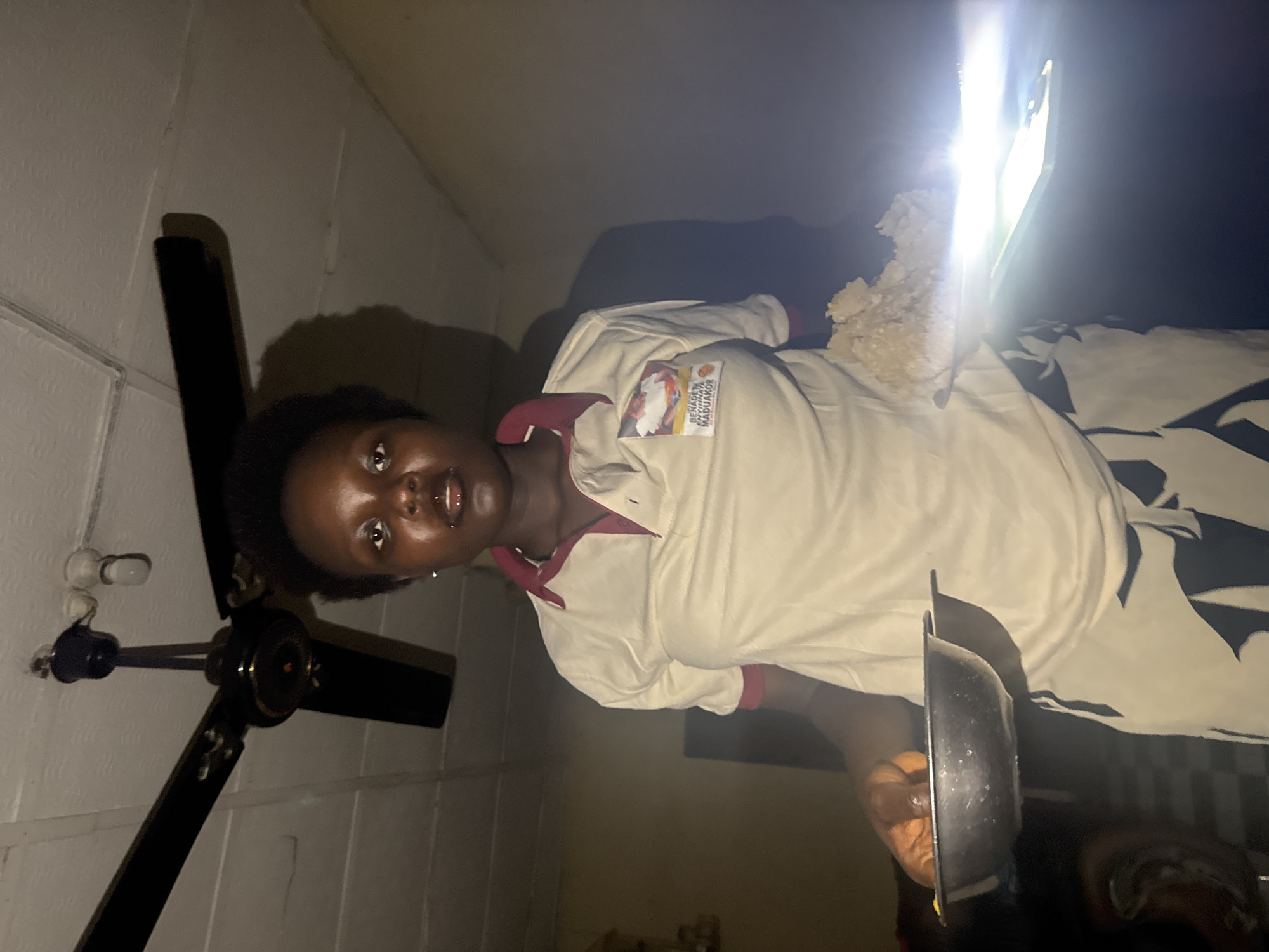 A person with dark skin and short, dark hair is holding a dark, round pan and an illuminated phone displaying white material in what appears to be an indoor setting in Onitsha, Nigeria. The person wears a light-colored shirt with maroon trim and a logo on the upper left. A ceiling fan with black blades is mounted on the white, paneled wall. A lightbulb can be seen adjacent to the fan. The area is dimly lit. A patterned blanket is visible behind the person.