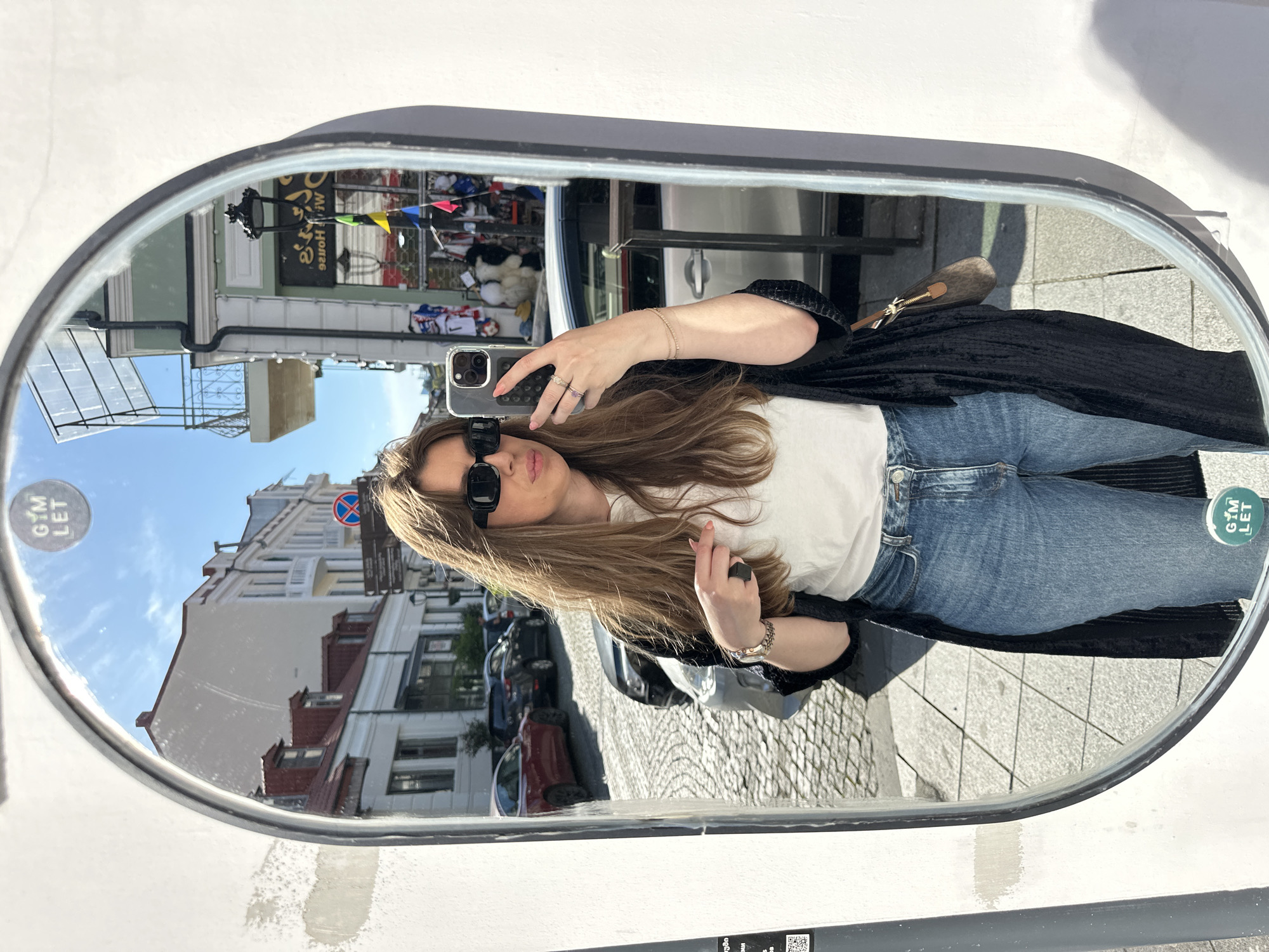 A woman is taking a selfie in a mirror mounted on a white wall. The mirror's reflection shows a street scene in Batumi, Georgia, with buildings, cars, and pedestrians visible. The woman is wearing jeans, a white t-shirt, a dark jacket, and sunglasses. She is holding a smartphone in her right hand. The reflection also shows a street with cobblestones and a building with a sign above a storefront.