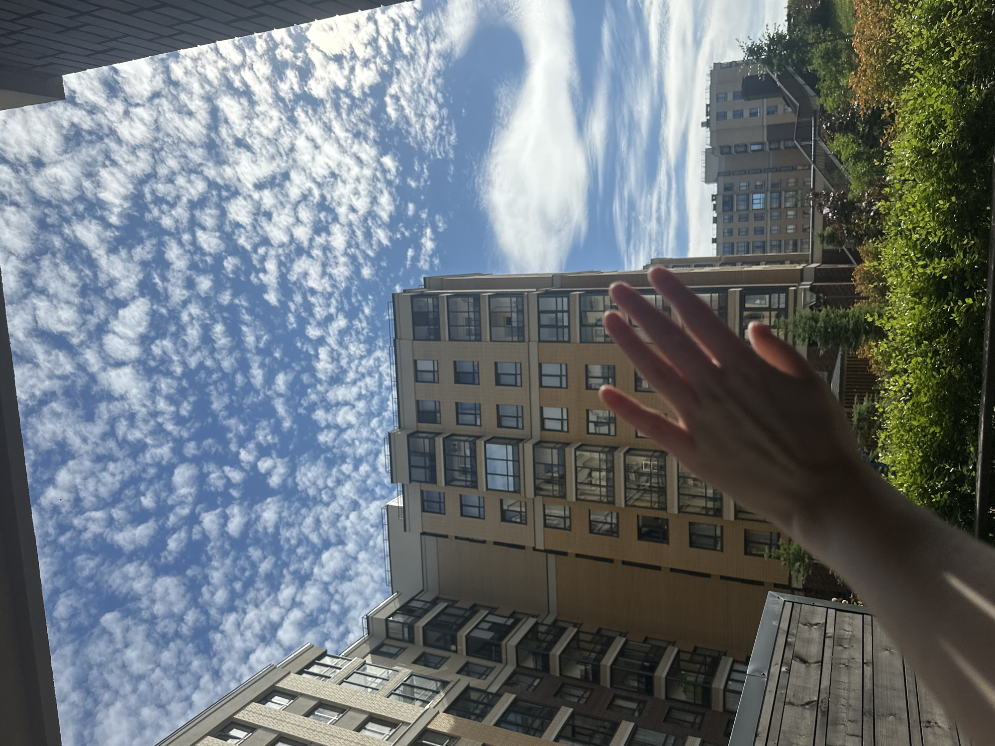 In an urban outdoor setting under a bright blue sky dotted with small, puffy white clouds, a hand is extended into the foreground. The hand is positioned to obscure a portion of the view. Behind the hand, a multi-story apartment building with a light brown facade and numerous windows dominates the mid-ground. To the left of this building, another apartment building, partially visible, has a darker facade. In the background, a second apartment building with a lighter facade and a visible staircase leading up to it is present. Dense green foliage and smaller trees are visible at the base of the buildings, suggesting a residential area with landscaping. The overall impression is a sunny day in a modern city, with the hand adding a human element to the scene.