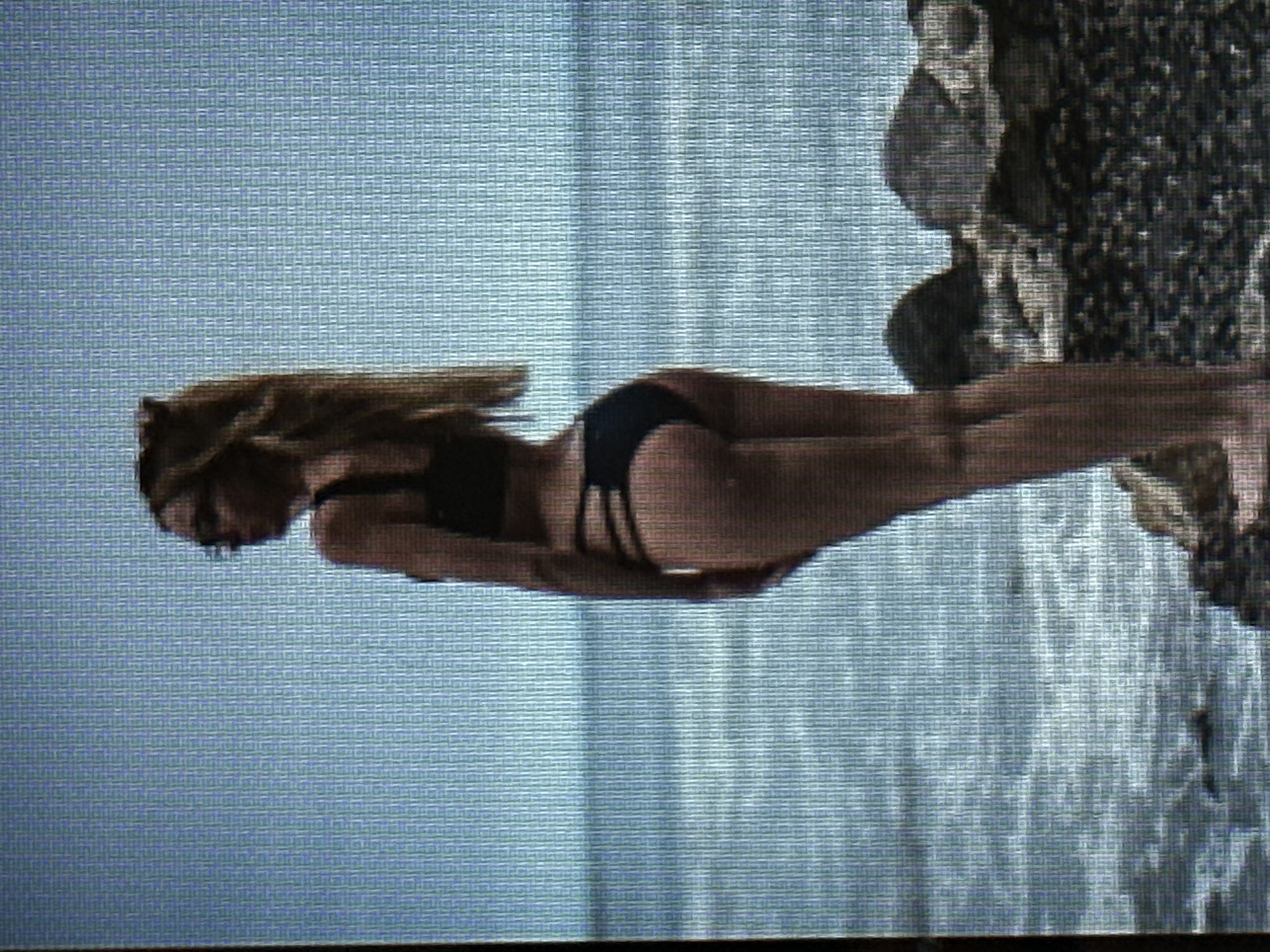 A full-body, side-view, portrait orientation photograph shows a woman from the shoulders down. The woman is wearing a black bikini and glasses. She is standing on a rocky shore with the ocean in the background. The sky is a pale blue. The image has a low resolution and appears to be a screen capture. There is no indication that this image is located in the city of Sofia, Bulgaria.