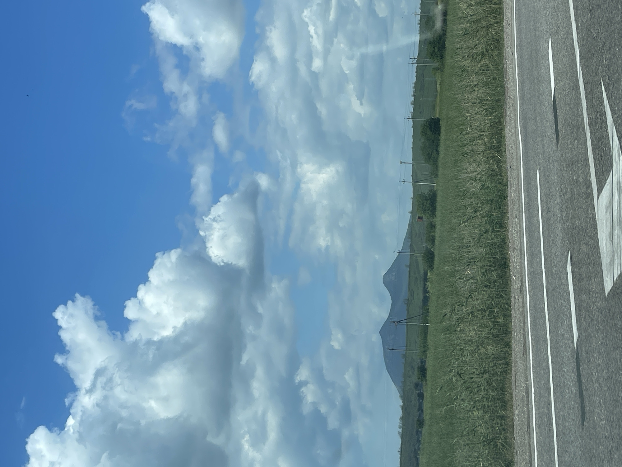The image shows a wide, asphalt road in the foreground with white lane markings. Beyond the road is a vast field of tall, green grass. In the distance, two prominent, rounded hills dominate the landscape, with a few scattered power poles and sparse vegetation on their slopes. The sky is a bright blue, filled with large, fluffy white clouds, some of which cast shadows on the hills and field. The overall scene depicts a rural, open landscape under a dynamic sky. The location is identified as Загорский, Russia.