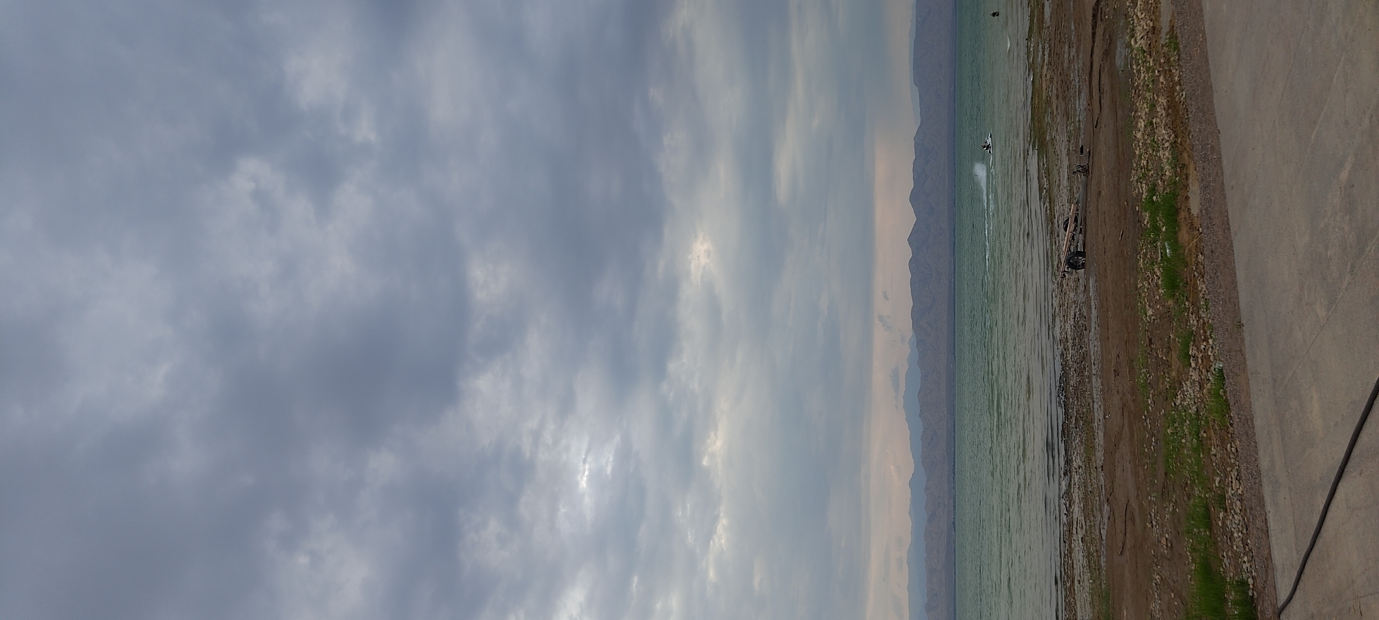 The media file shows a body of water, likely a lake or sea, with a distant mountainous landscape. The sky is overcast with dramatic, grey clouds, suggesting a possibly stormy or late afternoon/early morning atmosphere. On the water, a person is visible on a jet ski, creating a spray as they move. The shoreline is a mix of sand, pebbles, and some sparse green vegetation, with a paved area to the right of the frame. A trailer with wheels and some wooden planks is parked on the shore. The overall scene evokes a sense of a natural landscape with recreational activity. The location context suggests Chigdalik, Tajikistan.