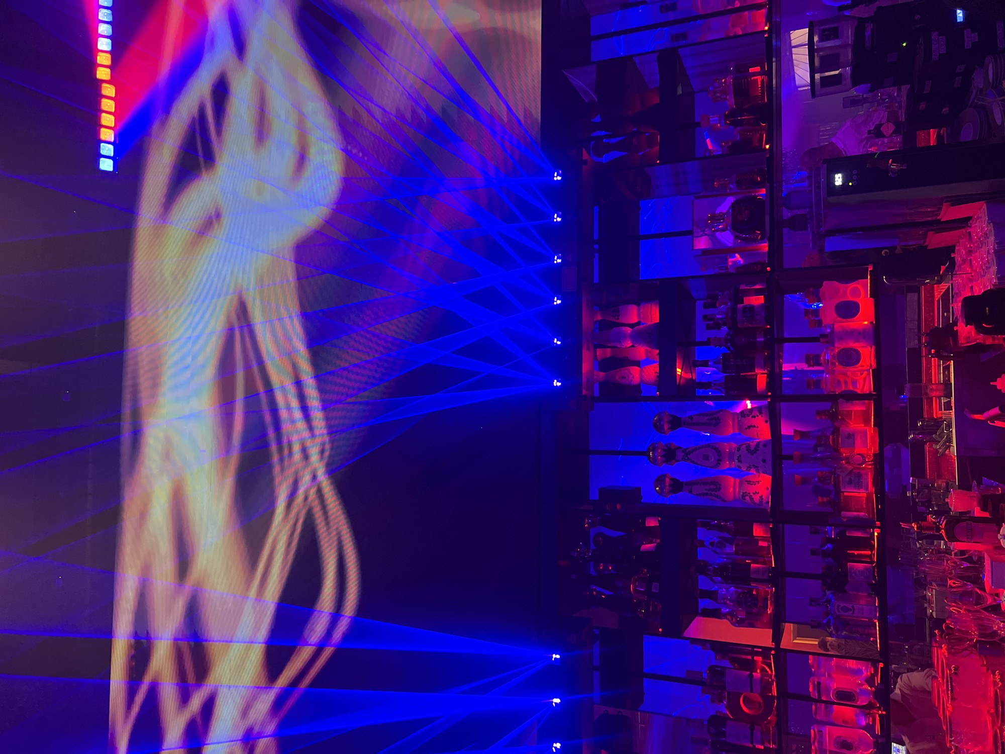 This image depicts a dimly lit bar or club interior. The upper portion of the scene is dominated by a large screen displaying an abstract light show with blue, pink, and yellow hues. Several blue laser beams are projected across the screen and into the space, creating a dynamic and energetic atmosphere. Below the screen, shelves are filled with numerous bottles of alcohol, illuminated by red and blue lights. Some of the bottles are tall and slender, while others are shorter and wider. Several distinctive tequila bottles, with ornate designs and stoppers, are prominent on the top shelf. In the lower part of the image, a bar counter is visible, lined with glasses and more bottles. A person, possibly a bartender, is partially visible behind the counter, suggesting an active establishment. The overall impression is one of a vibrant nightlife venue, likely in Choeng Thale, Thailand, given the context provided.