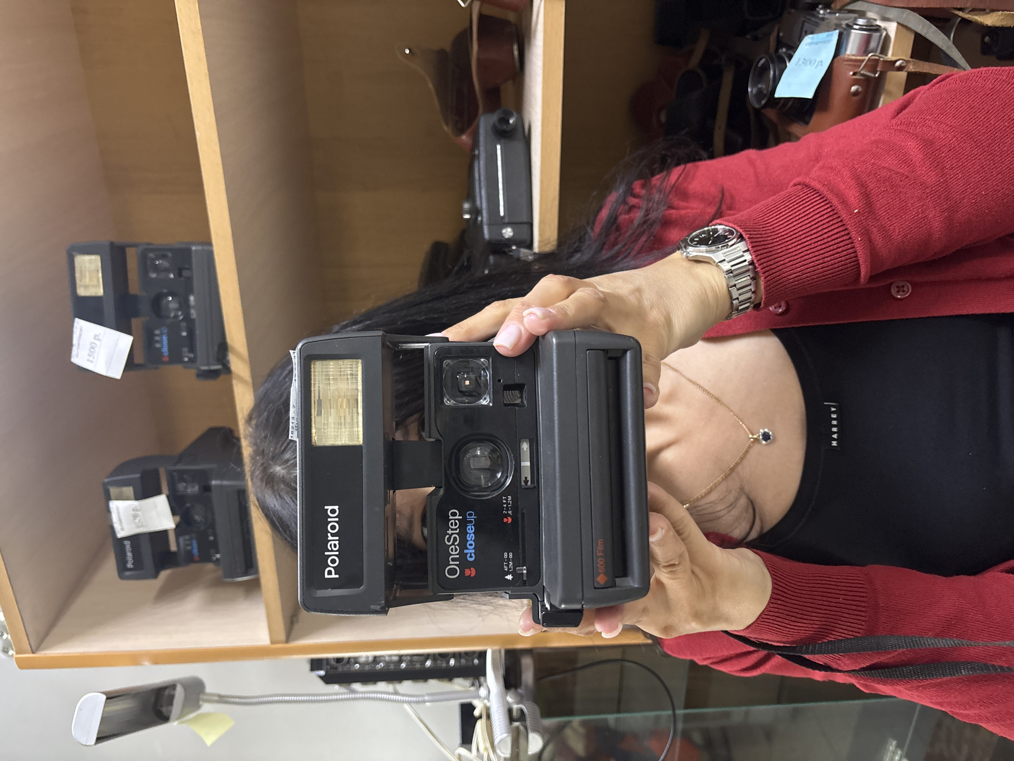 A close-up, upright portrait-oriented photo depicts a person holding a black Polaroid OneStep CloseUp instant camera, as if posing for or about to take a picture. The camera largely obscures the person's face, revealing only their chin, neck, and long dark hair. They are dressed in a red cardigan over a black top, which features the brand name "HARREY." A silver-toned watch with a metal bracelet is worn on their left wrist, and a delicate gold necklace with a dark blue pendant adorns their chest.
The Polaroid camera is the central focus, clearly displaying "Polaroid," "OneStep closeup," "4FT-∞," "1.2M-∞," and "600 Film" text. The person's hands, with manicured nails, hold the camera steadily.
The background shows an indoor setting, likely a store or display area in Chaikovsky, Russia, with light wooden shelves. Several other vintage cameras and camera accessories are arranged on these shelves. Price tags are visible on some items, with one reading "1500 р." (rubles) and another "1300p." A flexible desk lamp can be seen in the lower left. The image suggests an engagement with vintage photography equipment.