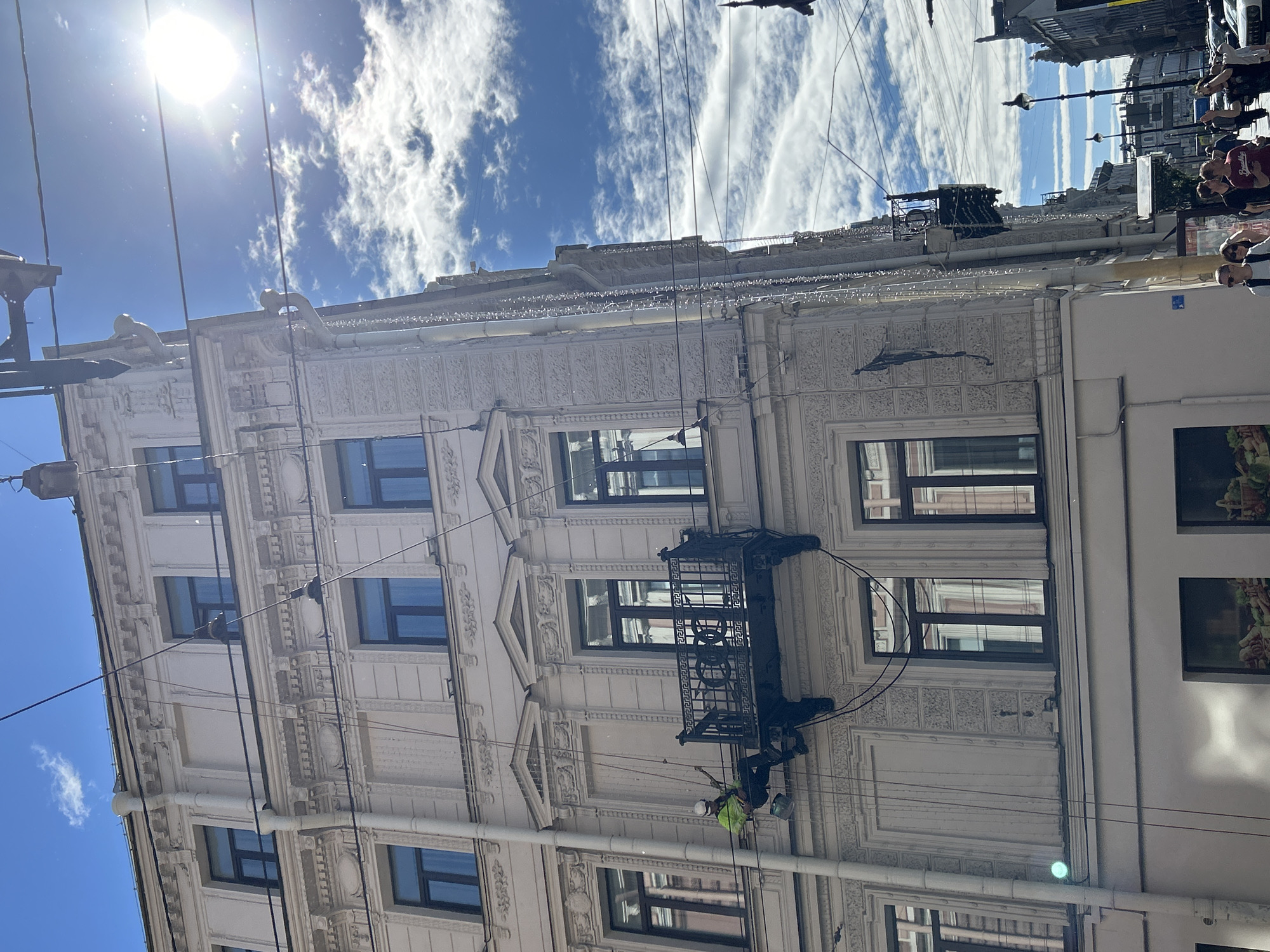 A person wearing a yellow jacket and a white helmet is suspended by ropes and working on the facade of a light-colored, ornate building in an urban environment. The person is on a small balcony-like platform and appears to be engaged in some form of maintenance or cleaning. Several windows with dark frames are visible on the building. Overhead, there are numerous electrical wires crisscrossing the bright blue sky, which is dotted with white clouds. Sunlight is strong and glinting off the wires and the building's surface. In the background, other buildings and streetlights are visible, as well as a street with cars and pedestrians. The scene appears to be a sunny day in a city.