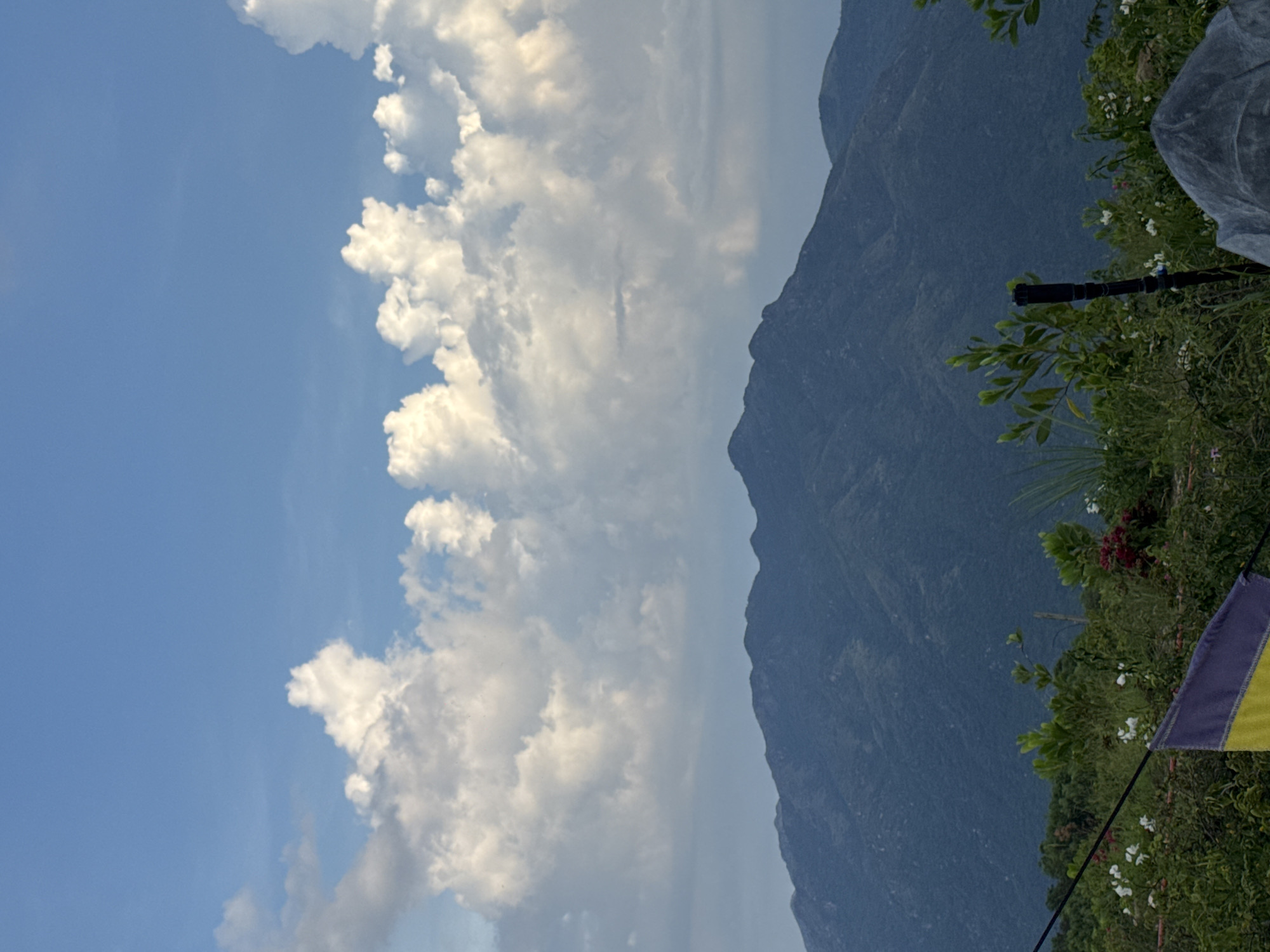 The image captures a scenic view of mountains and a cloudy sky, likely taken from a vantage point in Diên Khánh, Vietnam. In the foreground, lush green vegetation with scattered white and red flowers is visible. A dark pole-like object stands among the plants, and a portion of a flag, colored yellow and purple, is draped across the lower left corner. The middle ground features the dense, green slopes of a mountain range, stretching across the frame. The background is dominated by a vast blue sky filled with large, puffy white clouds, illuminated by sunlight. The overall atmosphere is tranquil and natural, suggesting an outdoor setting with a panoramic view.