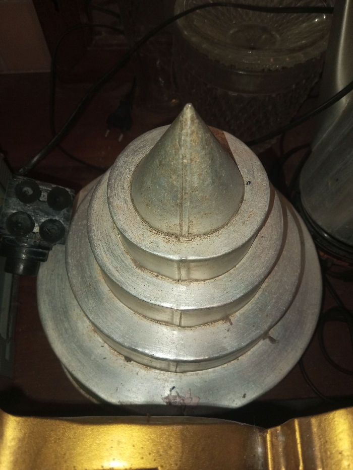 An overhead shot displays a tiered, conical metal object, resembling a retro-futuristic tower or decorative piece. The object is tarnished, showcasing age and wear. A black power cord and a black electrical component are visible to the left. A clear glass textured bowl sits in the background, along with another shiny metallic object. A glimpse of a gold, ornate item is also visible at the bottom.