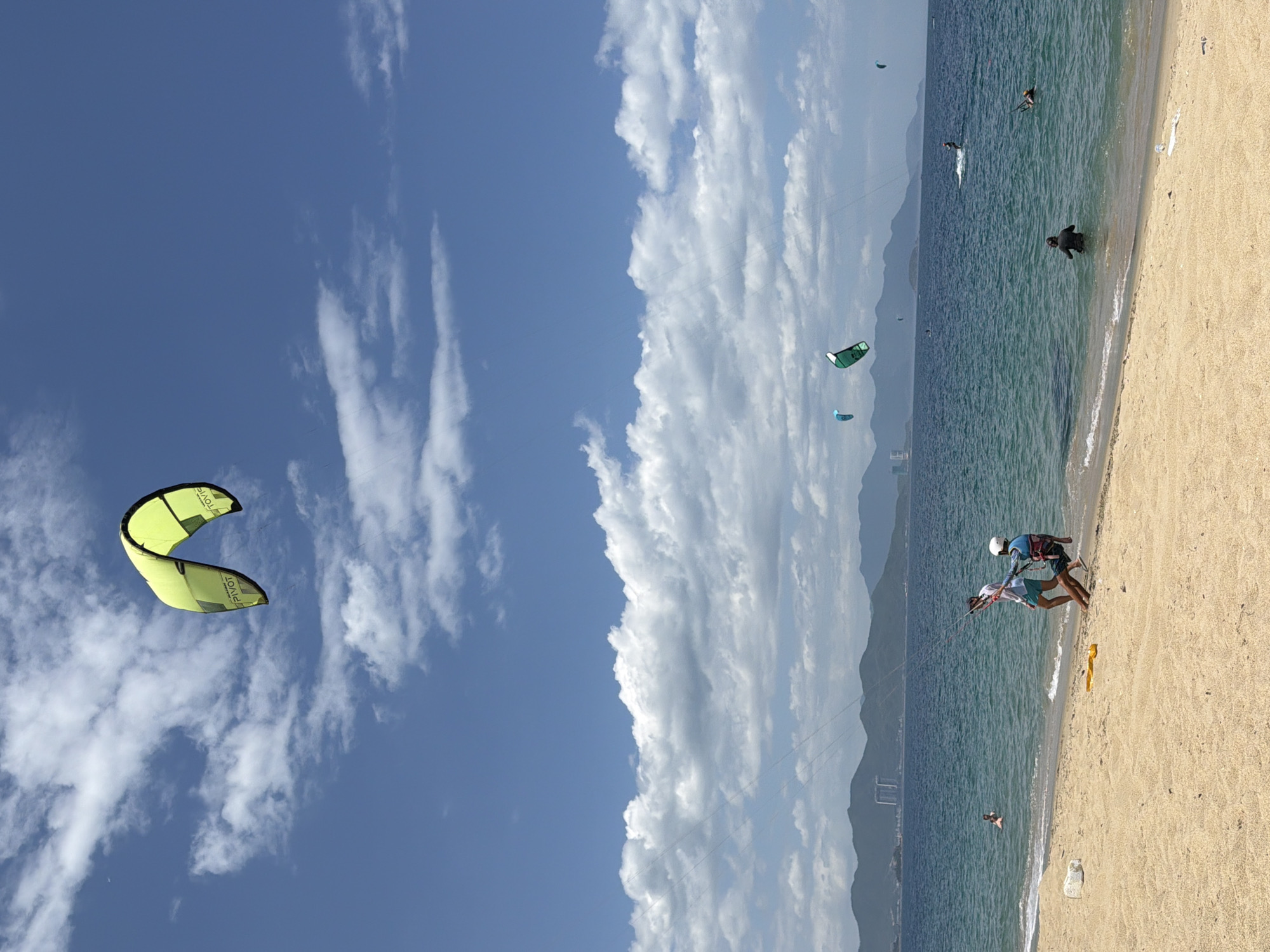 This image depicts a scenic beach in Nha Trang, Vietnam, featuring a clear blue sky dotted with white clouds. Several kitesurfers are visible in the azure ocean, engaging in their sport. On the sandy shore, two individuals are present; one is standing and appears to be instructing the other, who is seated and wearing a harness. The background shows a range of mountains and distant buildings, suggesting an urban coastline. The water is calm with gentle waves lapping the shore.
