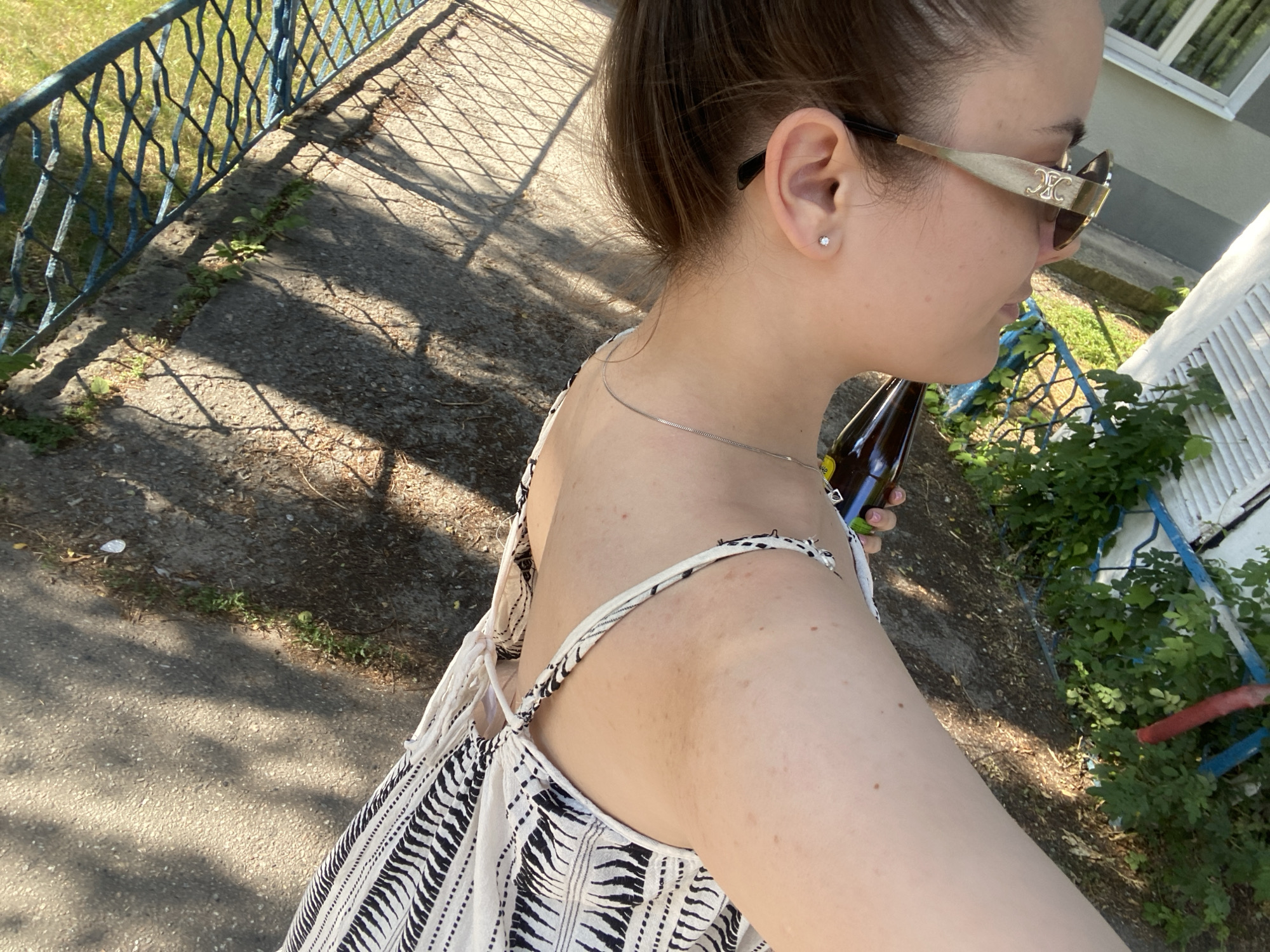 A woman, seen from the shoulder down, stands outside on a paved area near a blue metal fence. She wears a white and black patterned dress, sunglasses, and a silver necklace. She holds a dark glass bottle in her right hand. The ground is a mix of asphalt and dirt with some sparse vegetation. Sunlight casts shadows from the fence onto the ground. The setting appears to be an outdoor public space in Poltava, Ukraine.