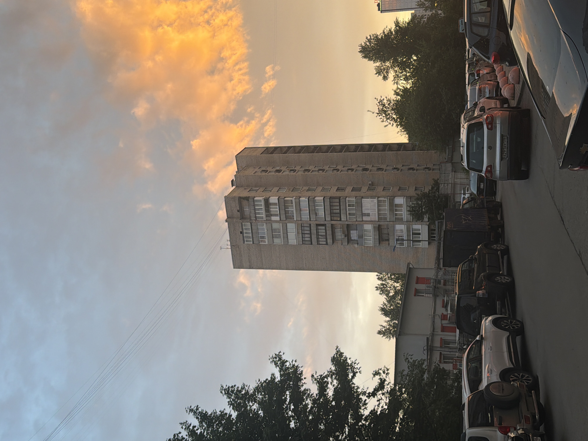 In an urban setting, a tall, brick apartment building stands on the right side of the image, illuminated by the warm glow of an orange sunset. The sky above is filled with clouds, some tinged with yellow and orange from the setting sun, while others are a soft blue. Electrical wires stretch across the sky. To the left of the apartment building, a dense green tree partially obscures the view. In the foreground, a street is lined with parked cars, including SUVs and a dark truck. A low-rise building with red-trimmed windows is visible behind the parked cars. The scene suggests a residential area at dusk.