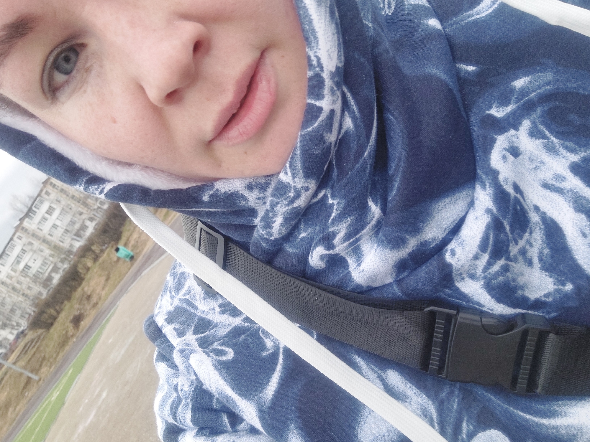 The image is a selfie of a person wearing a blue and white tie-dye hoodie with white drawstrings and a black strap with a black buckle across the chest. The person has fair skin, light-colored eyes, and their face is partially visible. In the background, there is a multi-story residential building. The location appears to be the city of Chaikovsky, Russia, based on visual cues consistent with the typical urban landscape and architecture found in Russian cities. The photo seems to be taken outdoors, possibly near a park or recreational area, with a glimpse of a green area in the background.