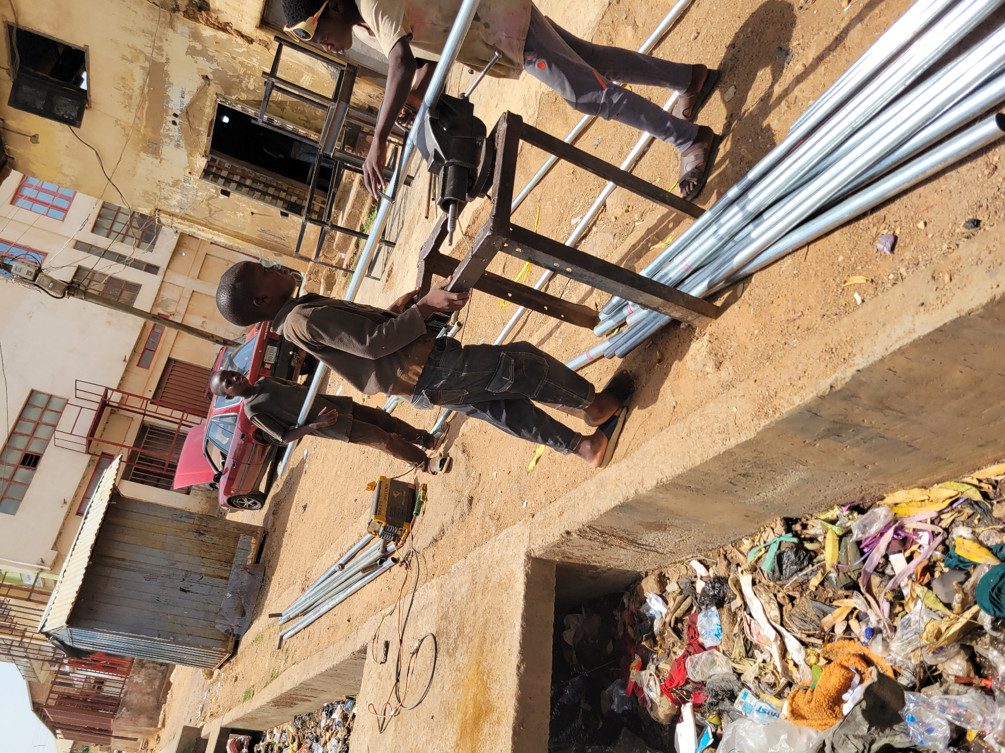 The image shows three African males in Jos, Nigeria, working on what appears to be a metal frame with a vise attached. Two of the males are actively engaged in manipulating the frame, while the third stands to the side. The foreground features a pile of metal pipes adjacent to the frame. A yellow electrical device, possibly a generator or inverter, sits nearby with wires leading to the workers. The background reveals a dilapidated urban setting with aged buildings and a red car parked near a corrugated metal structure. A concrete drainage ditch filled with debris and litter runs along the side, contrasting the work activity. The image conveys a scene of informal labor amid urban decay in Jos, Nigeria.