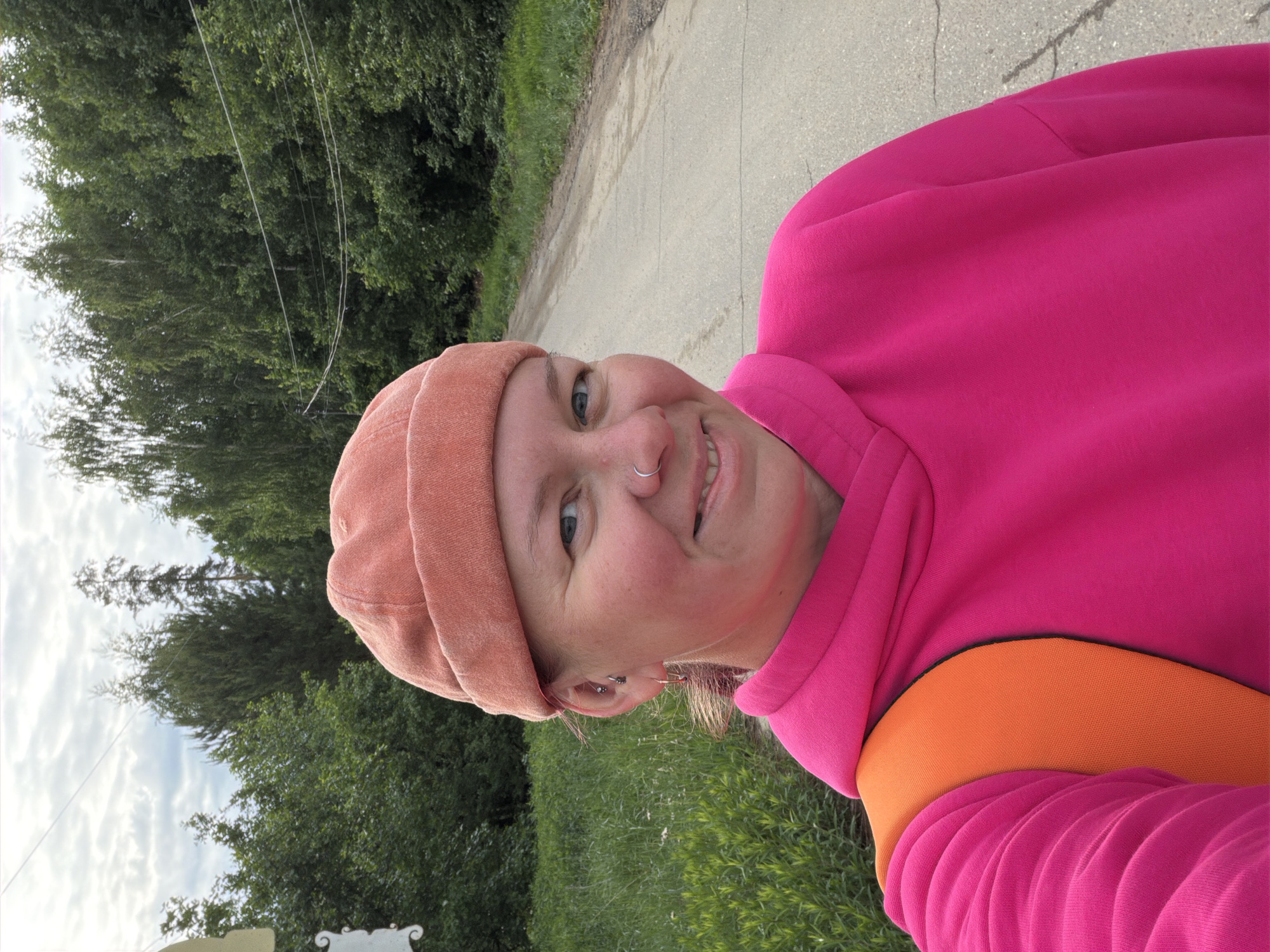 A selfie shows a smiling person in a pink hooded sweatshirt and a peach-colored beanie, standing on a paved road next to a grassy area and trees. The location is possibly in the city of Chaikovsky, Russia, although this cannot be confirmed from the image alone. The sky is cloudy. The person has a nose ring and visible ear piercings. An orange strap is visible under the sweatshirt.