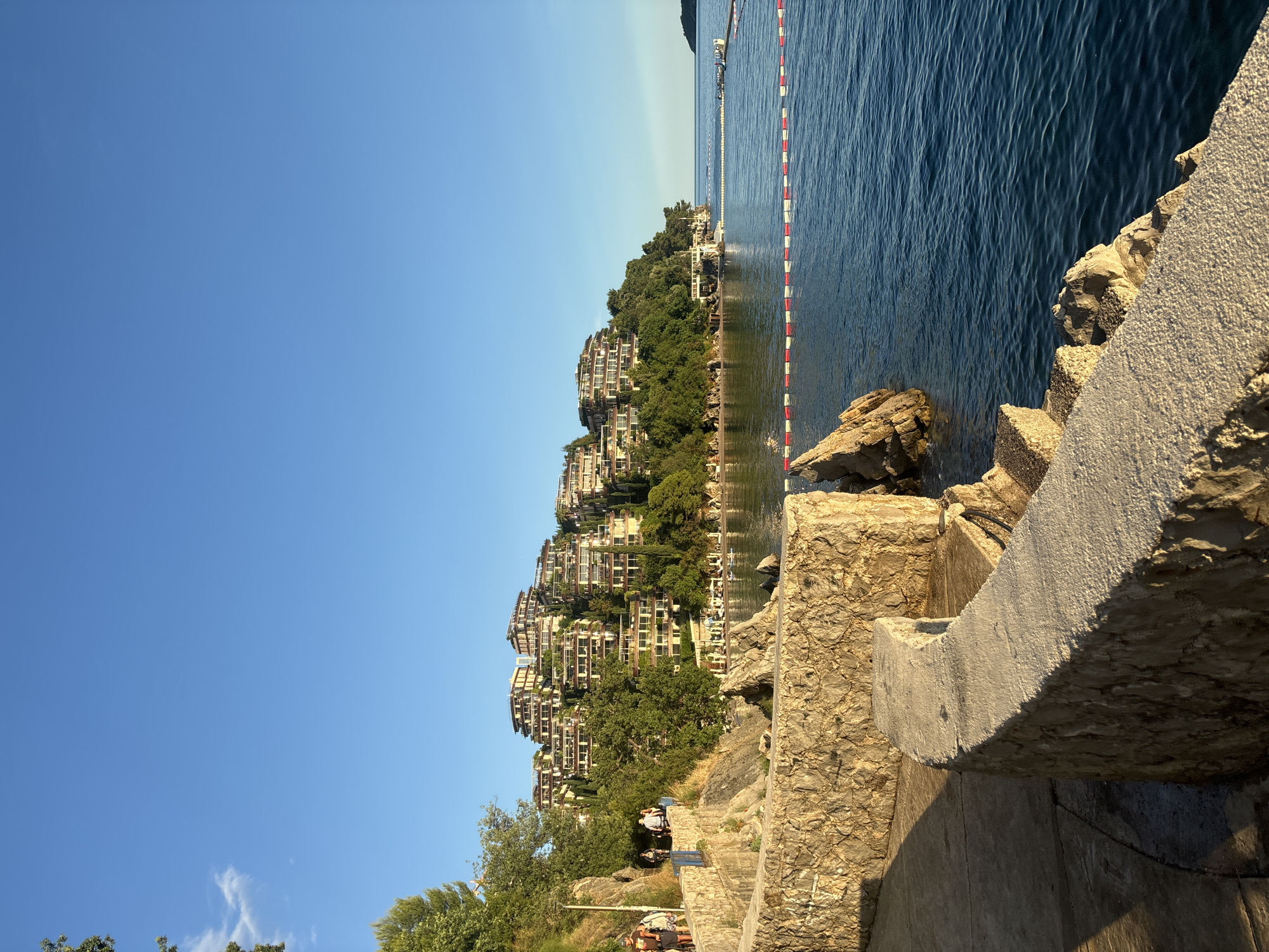 The image displays a vibrant coastal landscape under a clear, bright blue sky, characteristic of a Mediterranean setting. In the foreground, a rugged, rocky shoreline with textured concrete structures forms a path or viewing area, illuminated by direct sunlight that casts distinct shadows.
To the right, a calm, dark blue body of water, likely a sea or bay, extends towards the horizon. A distinctive red and white striped buoy line stretches horizontally across the water, parallel to the shore, suggesting a designated area for swimming or boating. Further along the water, a jetty or pier with a small structure is visible.
Across the water, a lush green hill rises steeply, densely covered with trees. Numerous multi-story buildings, likely apartments or hotels, are constructed into the hillside, arranged in terraced fashion amidst the foliage. These buildings feature multiple balconies and large windows, reflecting the sunlight.
A few individuals are visible on the rocky path in the mid-ground, casually walking or standing, their features indistinct due to distance.
The time of day appears to be late morning or early afternoon, with abundant sunshine and no clouds in the sky. The scene evokes a peaceful and scenic vacation atmosphere, consistent with a coastal resort area such as Budva, Montenegro. No discernible text is visible in the image.