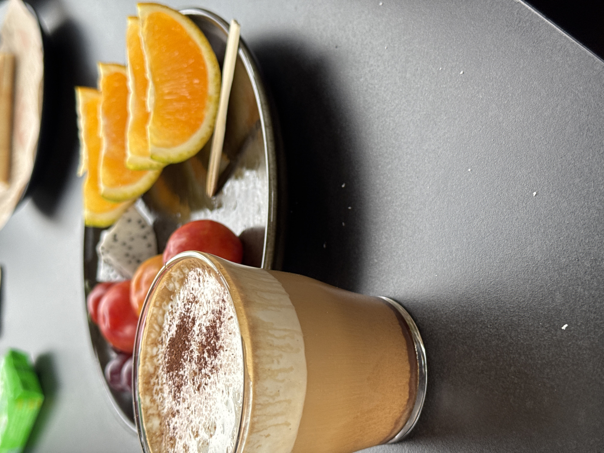 A high-angle, slightly rotated, and close-up shot reveals a still life composed of food and drink on a dark grey surface. A glass of layered coffee, topped with foamy cream and a dusting of brown powder, rests at the bottom. Adjoining this is a shallow, circular dish containing segmented oranges, other fruits, and a wooden stick. To the left, partial views of more food on a dish and a green item appear. There are no discernible people or interactions visible in the frame. The environment is defined by the flat, dark grey surface serving as the backdrop. There is no contextual information to determine this image’s location in Guilin, China.