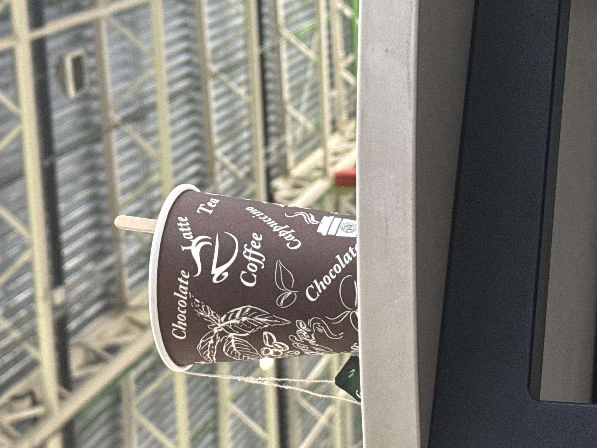 A close-up, rotated image captures a disposable coffee cup with a wooden stirrer, set against a blurred background of a large metal structure. The coffee cup is brown with white text and illustrations, including "Chocolate Latte," "Tea," "Cappuccino," "Coffee," and "Chocolat." Decorative patterns of coffee beans, leaves, and a steaming cup are also visible on its surface.
The cup is positioned to the left of a light-colored, vertically oriented surface, possibly concrete or stone, which runs along the right side of the frame and partially obscures the cup. To the right of this surface, a dark, granular-textured beam or frame is visible. A thin white string or thread hangs below the coffee cup, suggesting it might be suspended or tethered.
The background, rendered out of focus, reveals a complex network of interconnected metal beams, implying a large architectural or industrial structure, possibly scaffolding, a building under construction, or a bridge. The overall lighting indicates it is daytime. There are no individuals present in the scene, and no specific event or activity is taking place beyond the stationary presence of the coffee cup. The setting is in Lupanovo, Russia, suggesting an urban or industrial context for the backdrop.