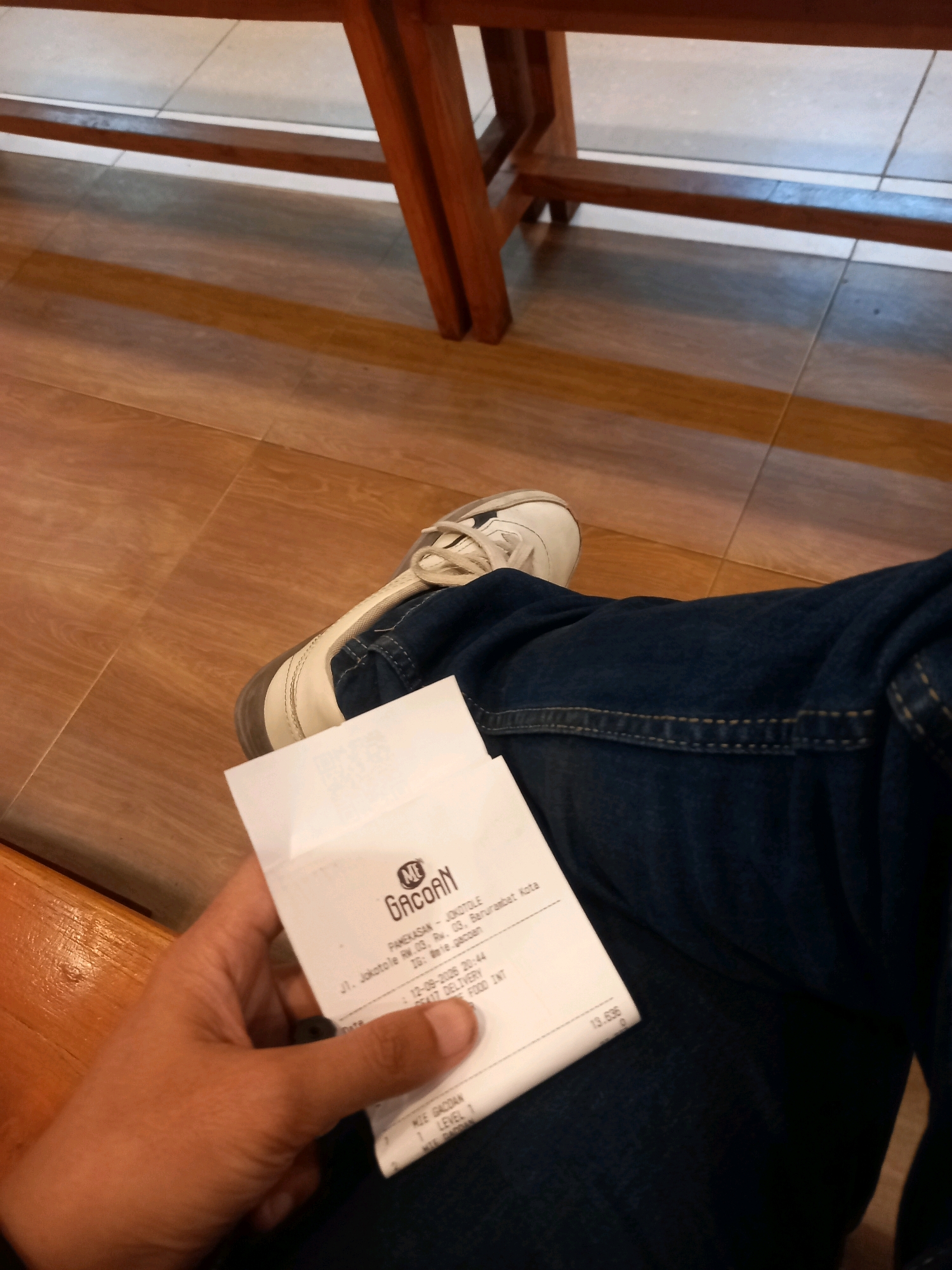 A person is holding a receipt from Gacoran restaurant in Pamekasan, Indonesia. The receipt shows the date and time of purchase as 12-09-2009, 20:44, and the total amount as 13,636. The restaurant's address is listed as Pamekasan RW.03, Rw. 03, Barurejamban, Kota Pamekasan, with the district of Barurejamban. The person is wearing blue jeans and white sneakers, and is seated on a wooden bench in what appears to be an indoor setting with tiled flooring.