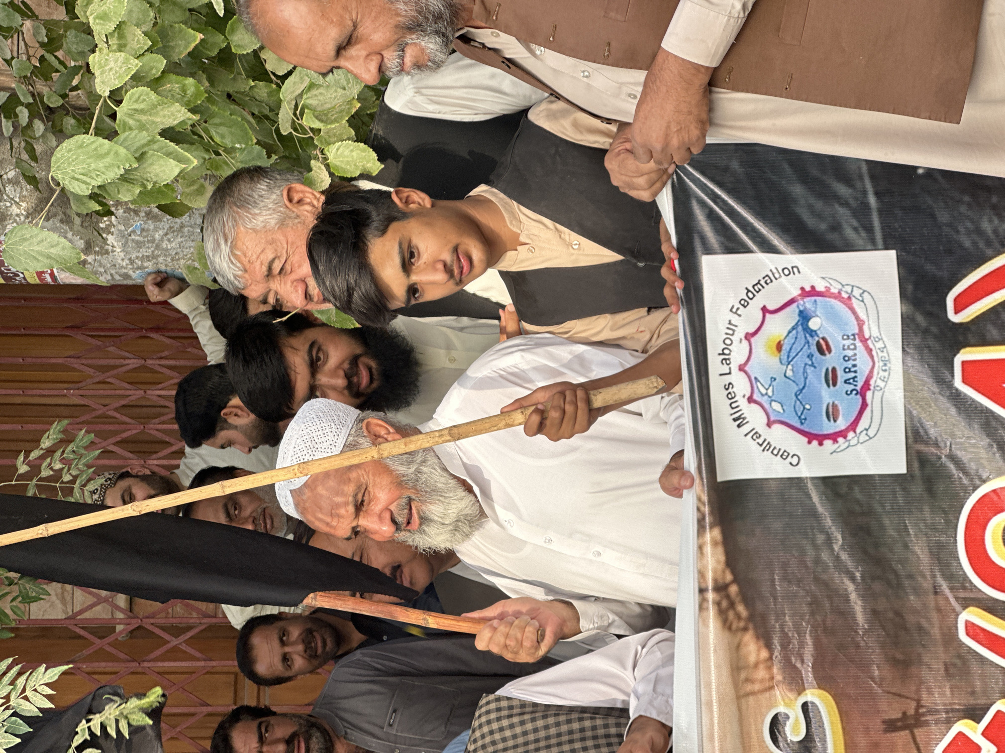The image depicts a group of men gathered outdoors, likely in the city of Quetta, Tehsil, Pakistan. In the foreground, an older man with a white beard and a white cap is holding a black flag attached to a wooden pole and another wooden stick. To his right, a young man wearing a dark vest over a beige shirt stands. To the far right, an older man with a beard, wearing a brown vest, is also visible. A banner with text and a logo is partially visible at the bottom of the image, showing the words "Central Mines Labour Federation" and a seal with a mountain and sun graphic. Behind the group, a reddish wooden gate and green foliage are visible, suggesting an outdoor setting. The lighting indicates it is daytime. The men appear to be participating in a protest or gathering.