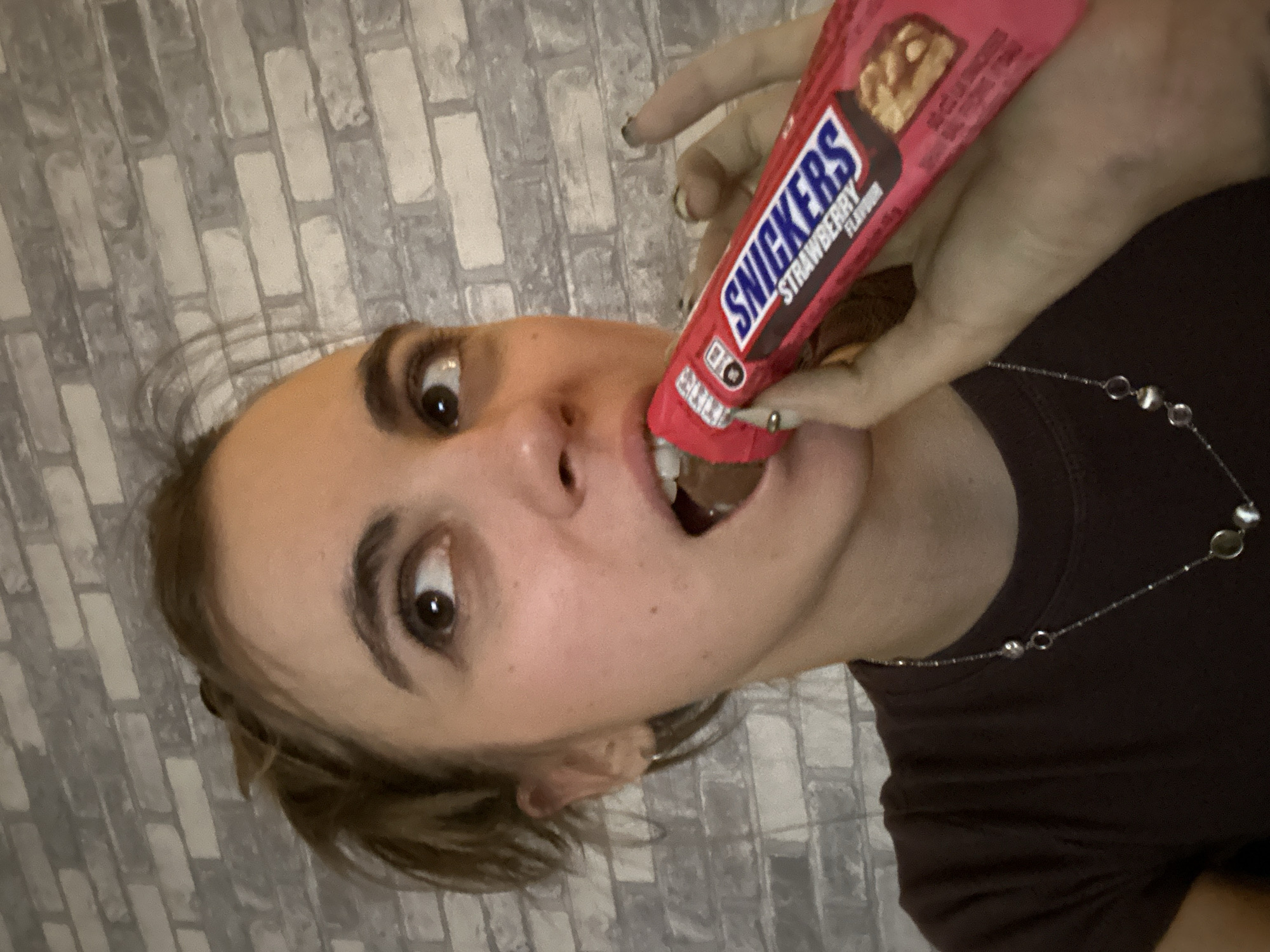 A woman is holding a strawberry flavored Snickers bar to her mouth as she prepares to take a bite. She is wearing a dark brown t-shirt and a silver necklace with round pendants. The background is a grey brick wall. The image is a close-up, focusing on the woman's face and the candy bar. The exact location in Moscow, Russia cannot be determined from the image.