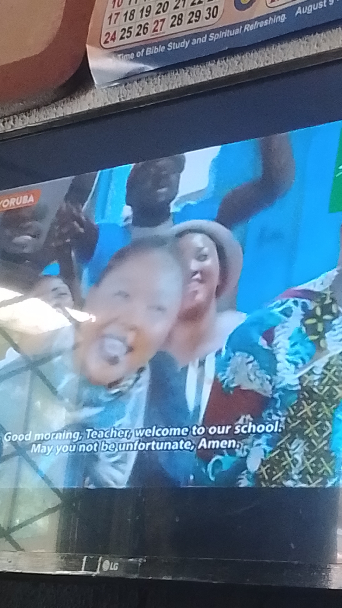 The image is a photograph of a television screen. The screen displays a scene with several people, including children and adults. The people appear to be gathered together, possibly for an event or a celebration. The background is a bright blue and shows what looks like a banner or a flag. There is text at the bottom of the screen that reads "Good morning, Teacher, welcome to our school. May you not be unfortunate, Amen." The television itself is black and has an LG logo on the bottom bezel. The image appears to be taken indoors, likely in a room with artificial lighting.
