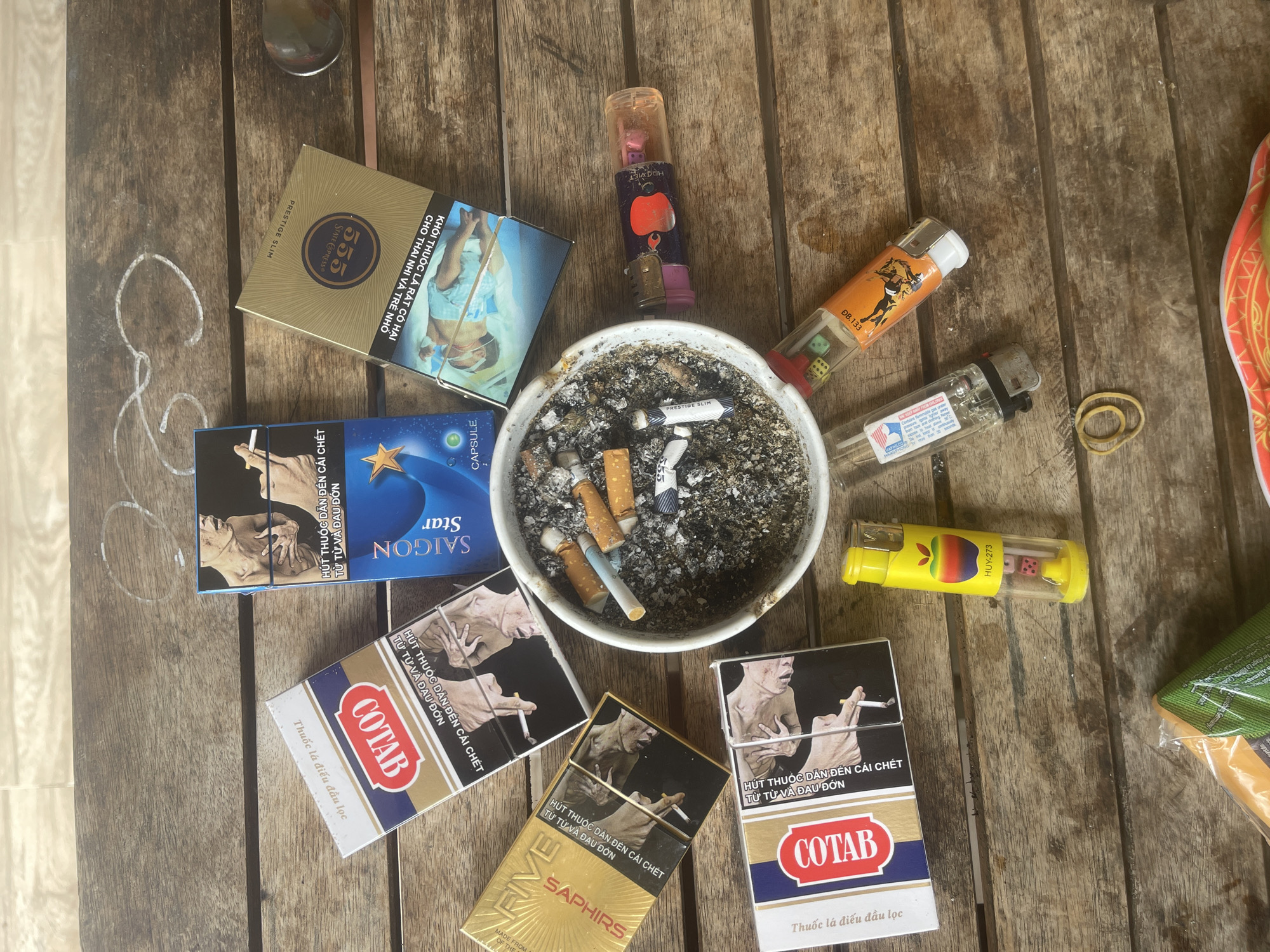 On a weathered wooden table, multiple packs of cigarettes are scattered, some open, revealing their contents. Among them are packs of "COTAB," "SAIGON Star," "555 State Express," and "FIVE SAPHIRES." Several cigarette butts are extinguished in a white ashtray. Three lighters are also present, one yellow with an apple design, and two clear lighters with colored liquids and what appear to be dice inside. A rubber band lies loose on the table. The scene is lit by natural light. This appears to be an outdoor setting, possibly in the city of Phan Thiet Ward, Vietnam, given the presence of local cigarette brands and the visual style.