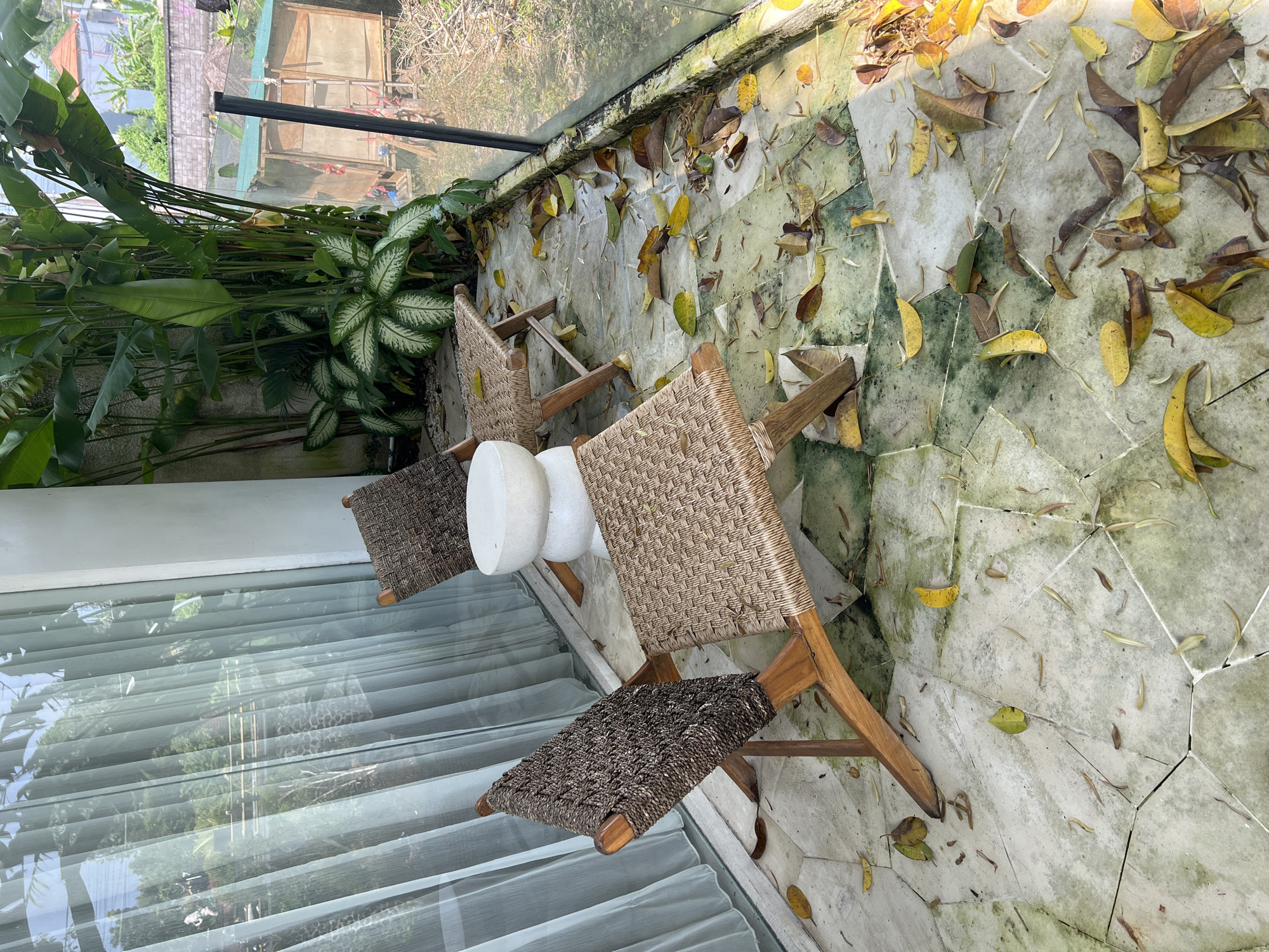 A high-angle view captures a balcony scene in Buduk, Indonesia. Three wooden chairs with woven seats surround a white cylindrical object, possibly a side table or stool. Fallen leaves are scattered on the patterned tile flooring. Lush greenery is visible on the left, contrasting with the urban structures seen through the balcony railing. A glass panel on the left reflects the surroundings, partially obscuring the view. The environment appears tranquil, suggesting a relaxed outdoor setting.
