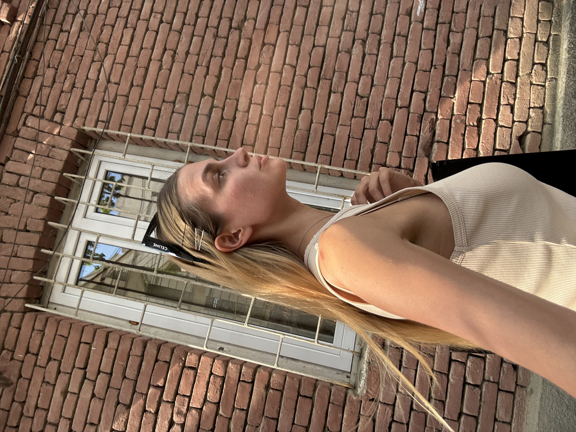 A woman with blonde hair is looking up, standing in front of a brick wall with a window. The window has bars. The location appears to be in Tbilisi, Georgia. She is wearing a light-colored tank top and has sunglasses resting on her head, held in place by hairpins.