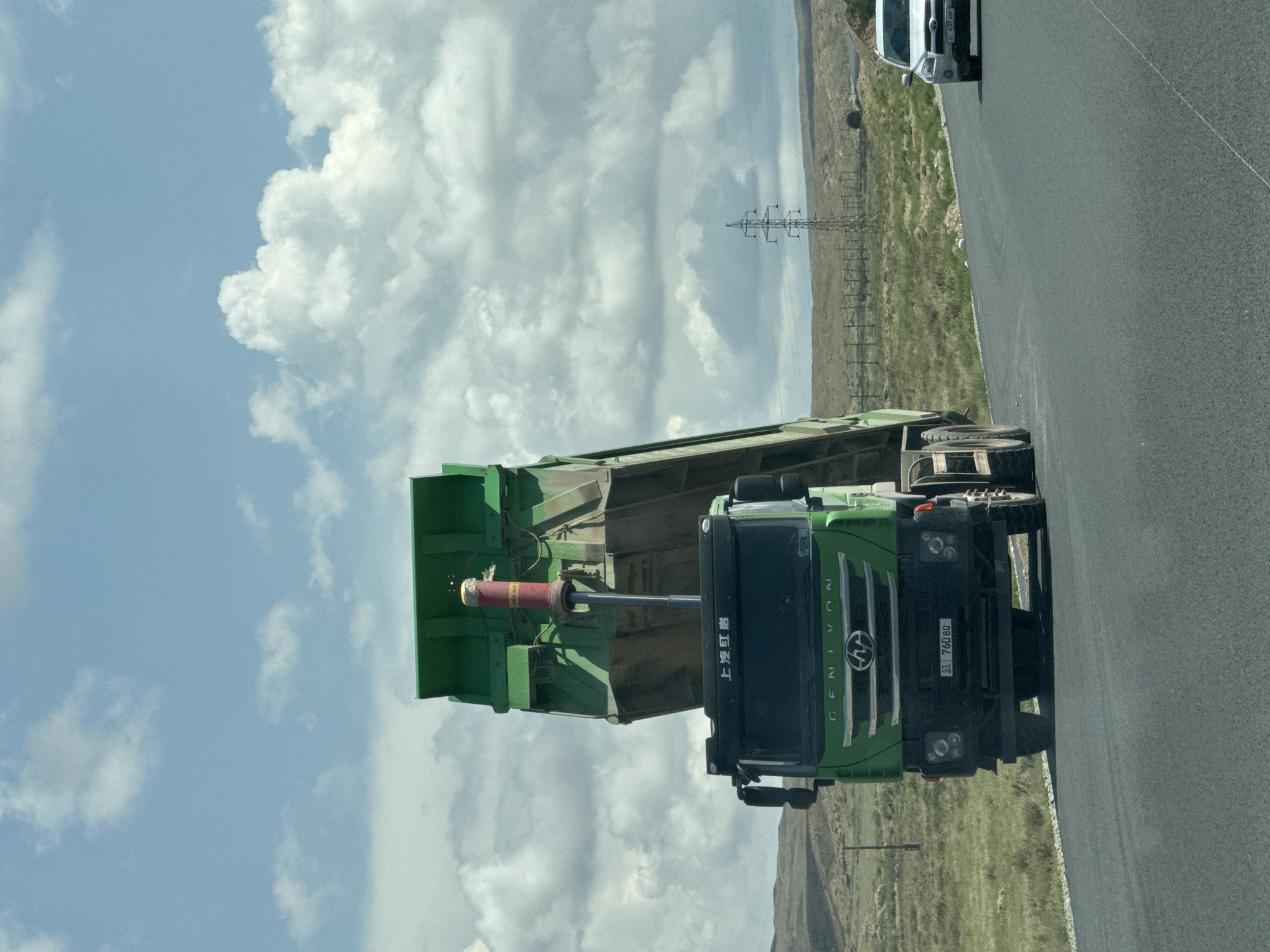 A green dump truck is parked on the side of a road with its bed raised. The truck has a license plate that reads "01 760 BQ." To the right of the truck, a white SUV is driving away. The scene is set in a rural landscape with rolling hills and a cloudy sky. A power line tower is visible in the distance. The road appears to be newly paved. The image was taken in the city of Kyzyl-Tuu, Kyrgyzstan.