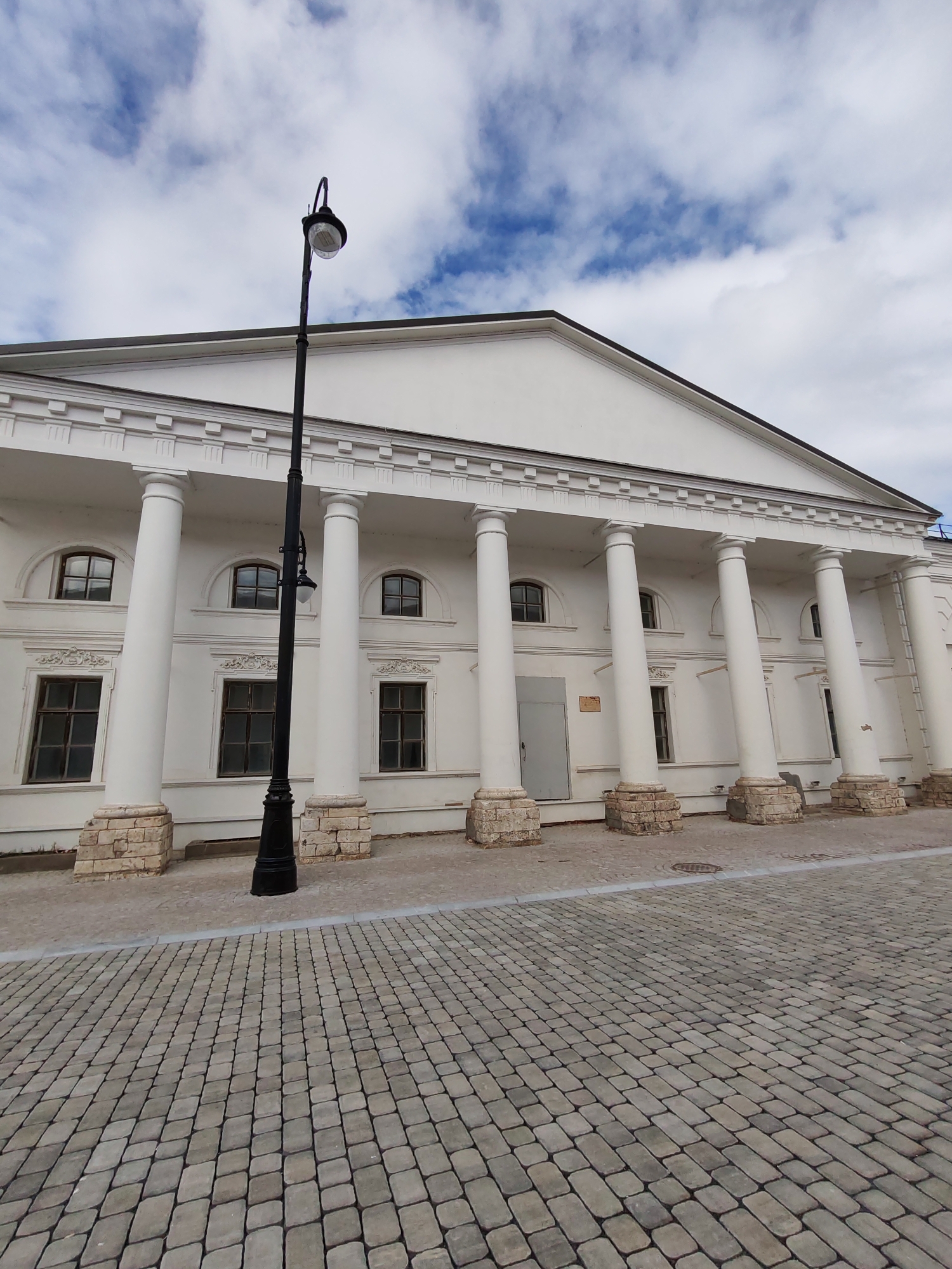 This media file captures a frontal view of a white, classical-style building with a portico supported by large, white columns. The building has a triangular pediment at the top, adorned with dentil molding. The facade features multiple windows, some arched and some rectangular, all with dark frames. The base of the columns rests on square, stone plinths. A black lamppost stands prominently in front of the building, slightly to the left of the center. The ground in front of the building is paved with interlocking grey cobblestones, with a lighter paved pathway leading up to the building.
The sky above is partly cloudy, with patches of blue sky visible between white cumulus clouds, suggesting it is daytime. The lighting indicates it is likely late morning or early afternoon. There are no people visible in the image, and no specific activity is taking place; the scene appears to be a static architectural view. The location is identified as Bol'shie Mkhi, Russia. No visible text is present on the building or in the immediate surroundings. The overall impression is one of historical architecture under a bright, partly cloudy sky.