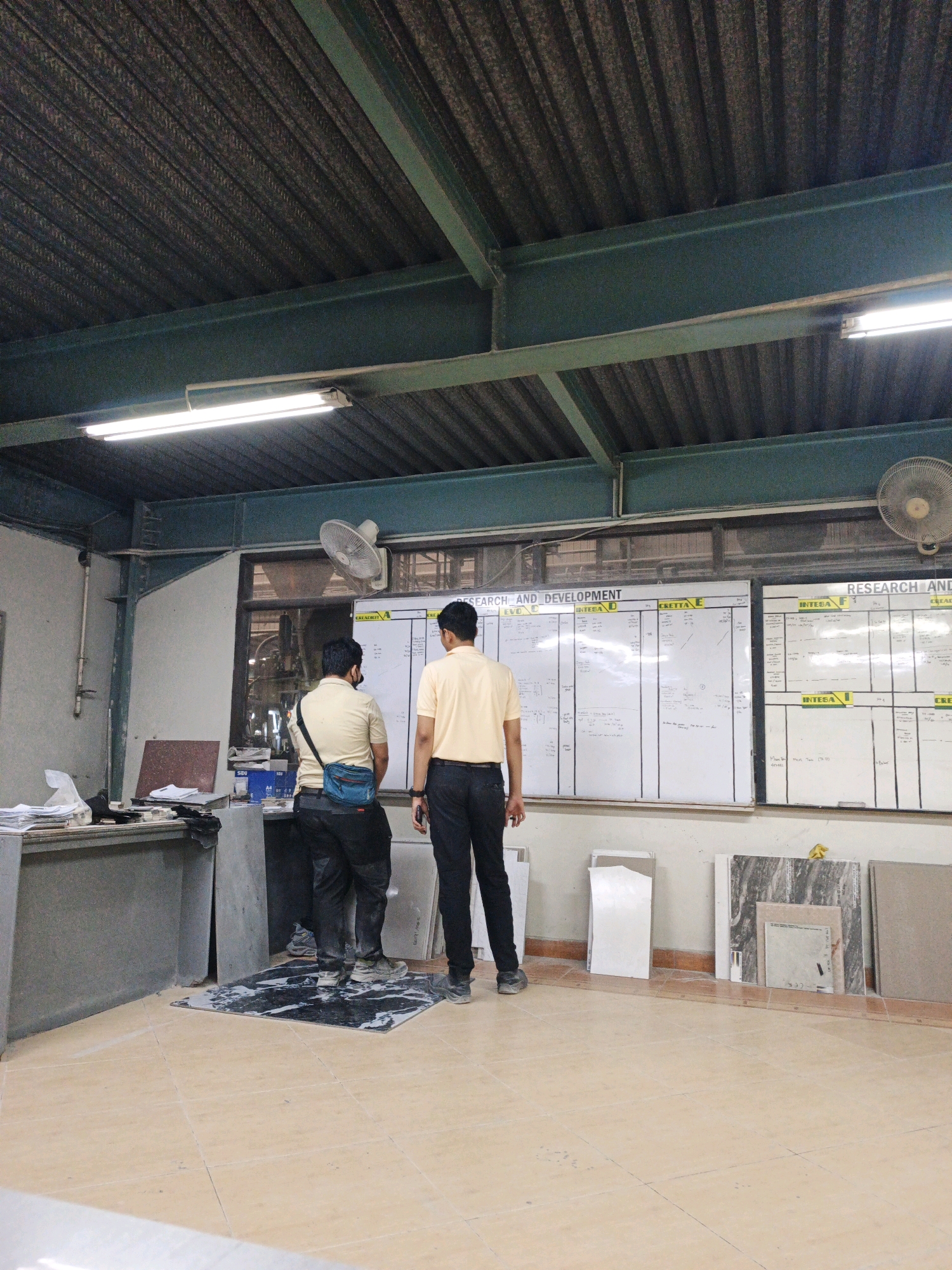The image depicts an indoor industrial setting, possibly a factory or workshop, located in Ngoro, Indonesia. Two men, both wearing light-colored shirts and dark pants, stand facing a large whiteboard covered in text and diagrams. One man is positioned slightly to the left and appears to be wearing a mask around his neck. The other man is standing to his right, closer to the whiteboard.
The room has a corrugated metal ceiling supported by dark green beams. Fluorescent lighting fixtures are mounted on the ceiling, illuminating the space. A ceiling fan is visible in the upper right portion of the image. To the left, there is a desk with papers and office supplies, and against the wall, a collection of large stone or tile samples are displayed. The floor is tiled in a beige color, with a dark patterned mat placed in front of the whiteboard. The overall environment suggests an active workspace where planning or review is taking place.