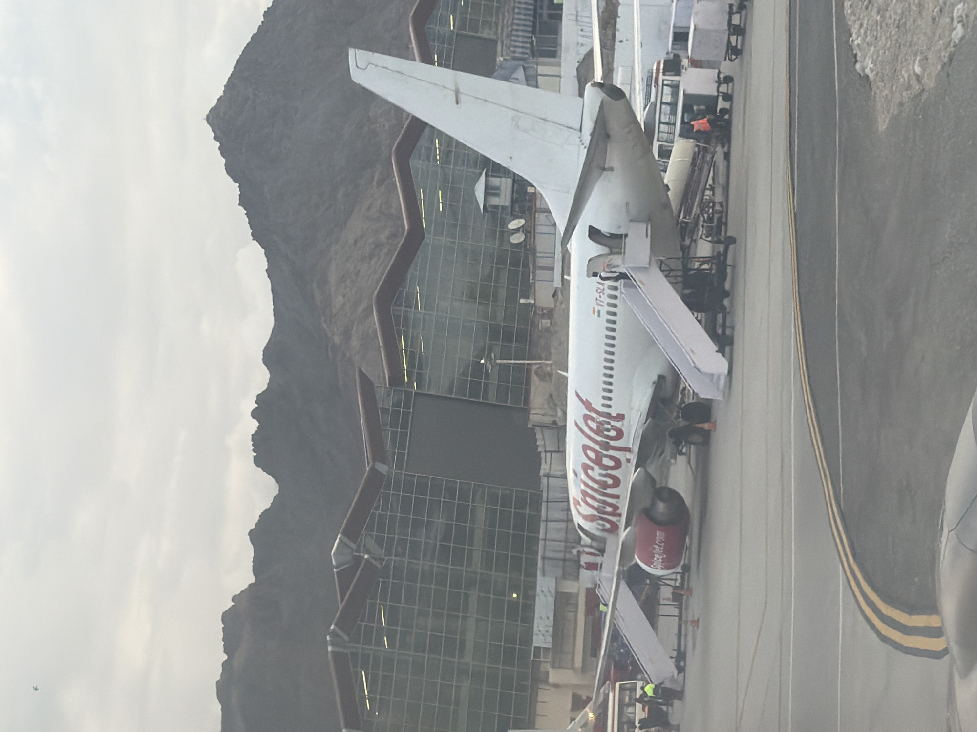 An airplane with the SpiceJet logo is parked on the tarmac of Leh Airport, India. The airport's modern glass facade terminal building is visible in the background, with rugged, mountainous terrain behind it. Several mobile stairs are connected to the aircraft for passengers. Airport personnel are visible near the plane, some of whom appear to be working with luggage carts and equipment. The sky is overcast, suggesting a potentially cool or hazy day.
