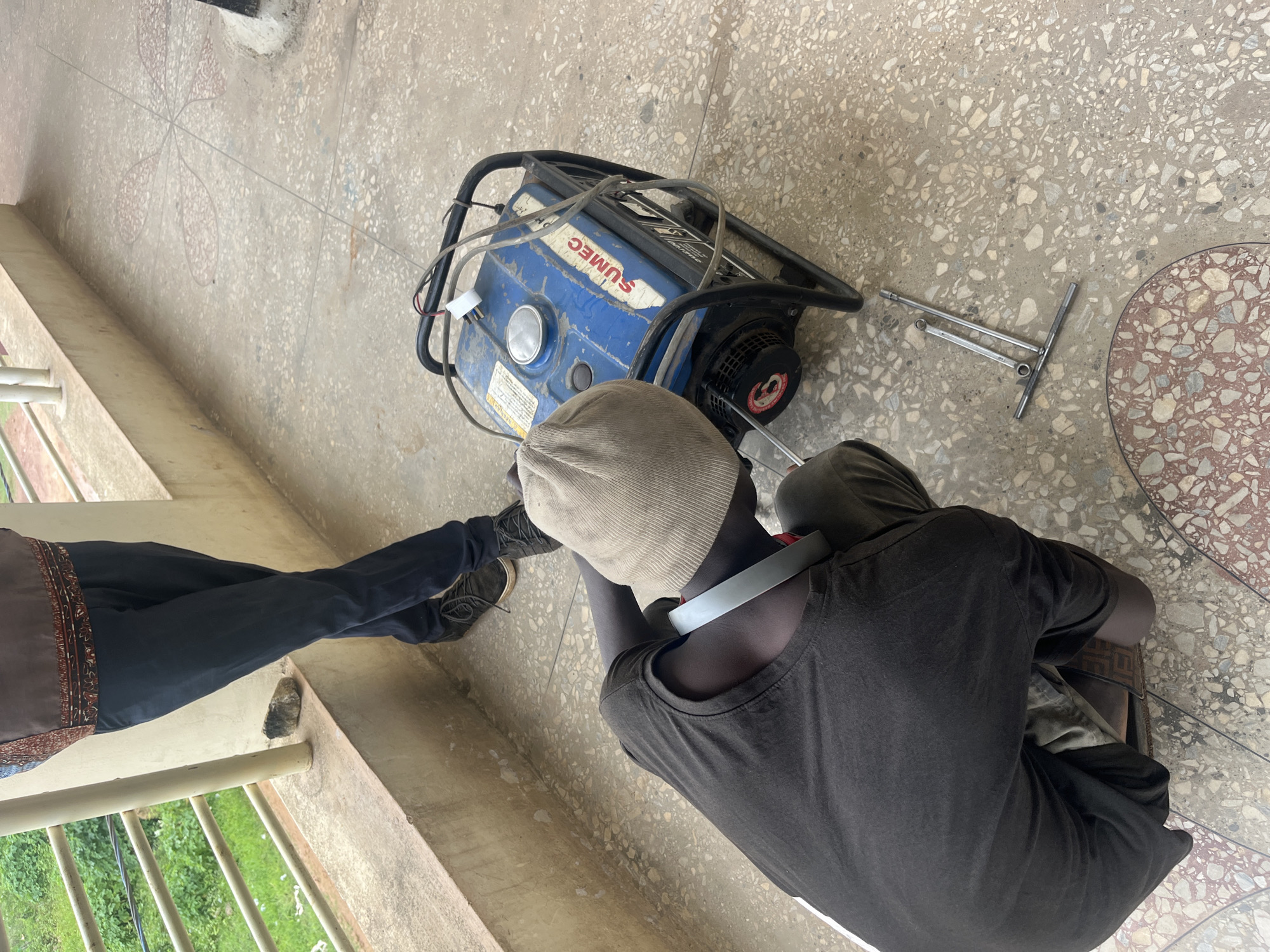 In Gura Top, Nigeria, a person wearing a brown and black patterned shirt and dark pants stands near a railing overlooking green vegetation. In the foreground, another person, wearing a dark t-shirt, brown pants, and a light brown knit cap, is crouched down, appearing to be working on a blue generator. The generator has "SUMEC" printed on its side and is surrounded by its metal frame. Tools, including a T-handle wrench and a socket wrench, are on the ground near the generator. The scene takes place on a terrazzo-tiled patio.