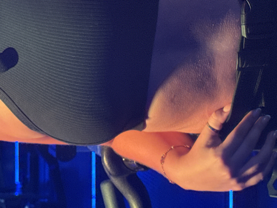 A close-up shows a person wearing a black, textured garment. Their arm, adorned with a delicate gold bracelet, rests on a gray handle. A ring is visible on their finger. The skin appears slightly damp. Blue light emanates from the background, possibly indicating an indoor setting. The overall composition suggests someone engaged in an activity or exercise.