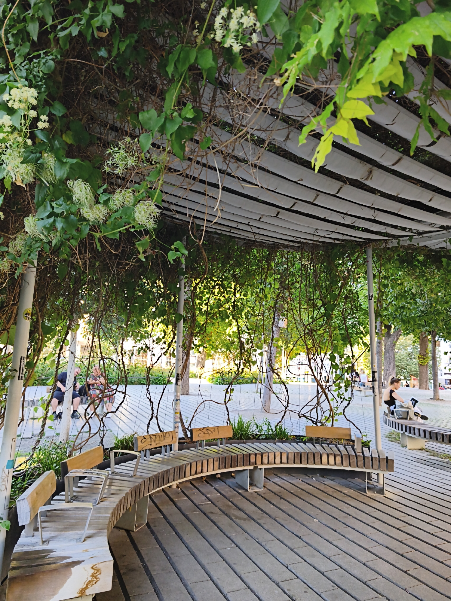 The image depicts a covered outdoor seating area with curved wooden benches under a canopy of green foliage and vines. The canopy is supported by metal poles and appears to be made of white fabric panels. The ground is paved with rectangular stones laid in a linear pattern. In the background, there are trees and a few people sitting on benches. One person, a woman wearing a black tank top, is seated on a bench with a laptop. Another couple, a man and a woman, are seated on a bench on the left side of the image, partially obscured by the vines. The scene suggests a public park or plaza, likely in a temperate climate given the lush greenery. This location appears to be the 'Stadtpark' in Vienna, Austria, a well-known urban park featuring similar modern outdoor seating and mature trees.