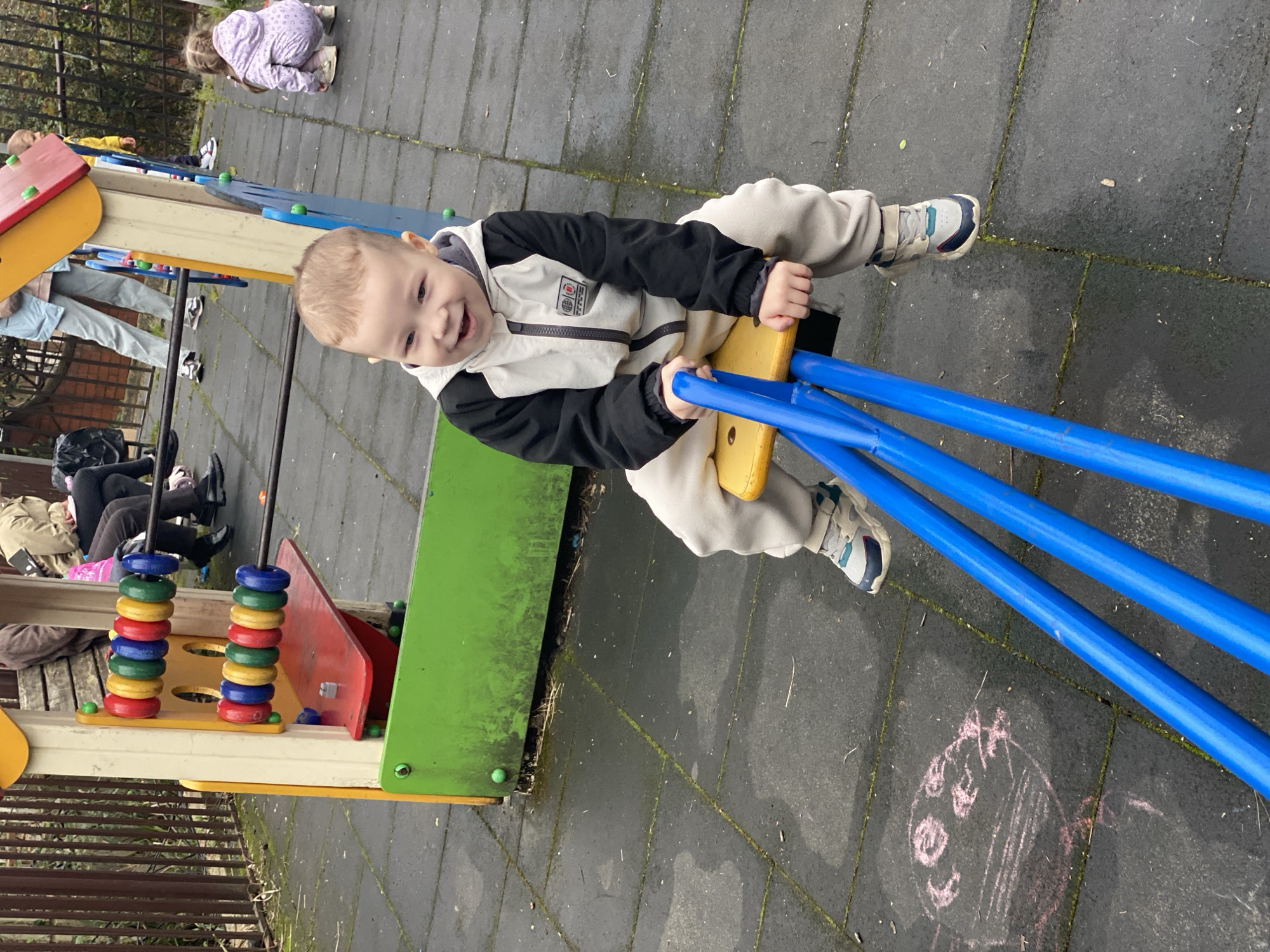 The image shows a young boy riding a seesaw in a playground. The boy is smiling and appears to be enjoying himself. The seesaw is blue and yellow. The playground is made of gray concrete and has other play equipment visible, including a structure with colorful rings. There are other children and adults in the background. The location appears to be outdoors, and the pavement has a wet look. There is an drawing in chalk of a face on the ground. There is no way to know from the image if the location is in Sochi, Russia.