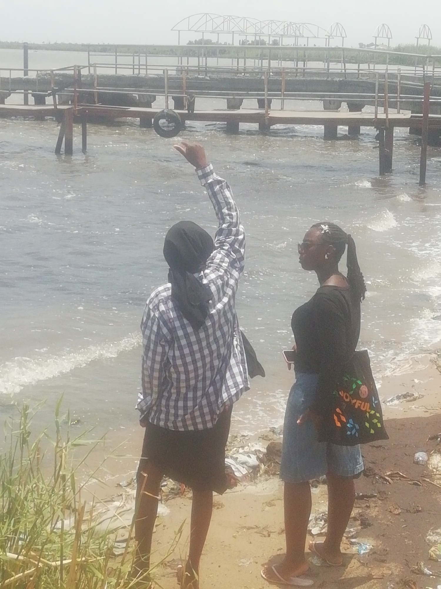 Two women stand at the edge of a body of water. One woman wears a plaid shirt, black skirt, and a headscarf, and has her arm raised in the air. The other woman wears a black top, denim skirt, sunglasses, and carries a black bag with "JOYFUL" printed on it. A rusted metal structure extends into the water. In the background, a bridge-like structure is visible. Trash litters the sandy bank. Tall grass grows in the foreground. The location appears to be a beach-like area in an undefined city and region.