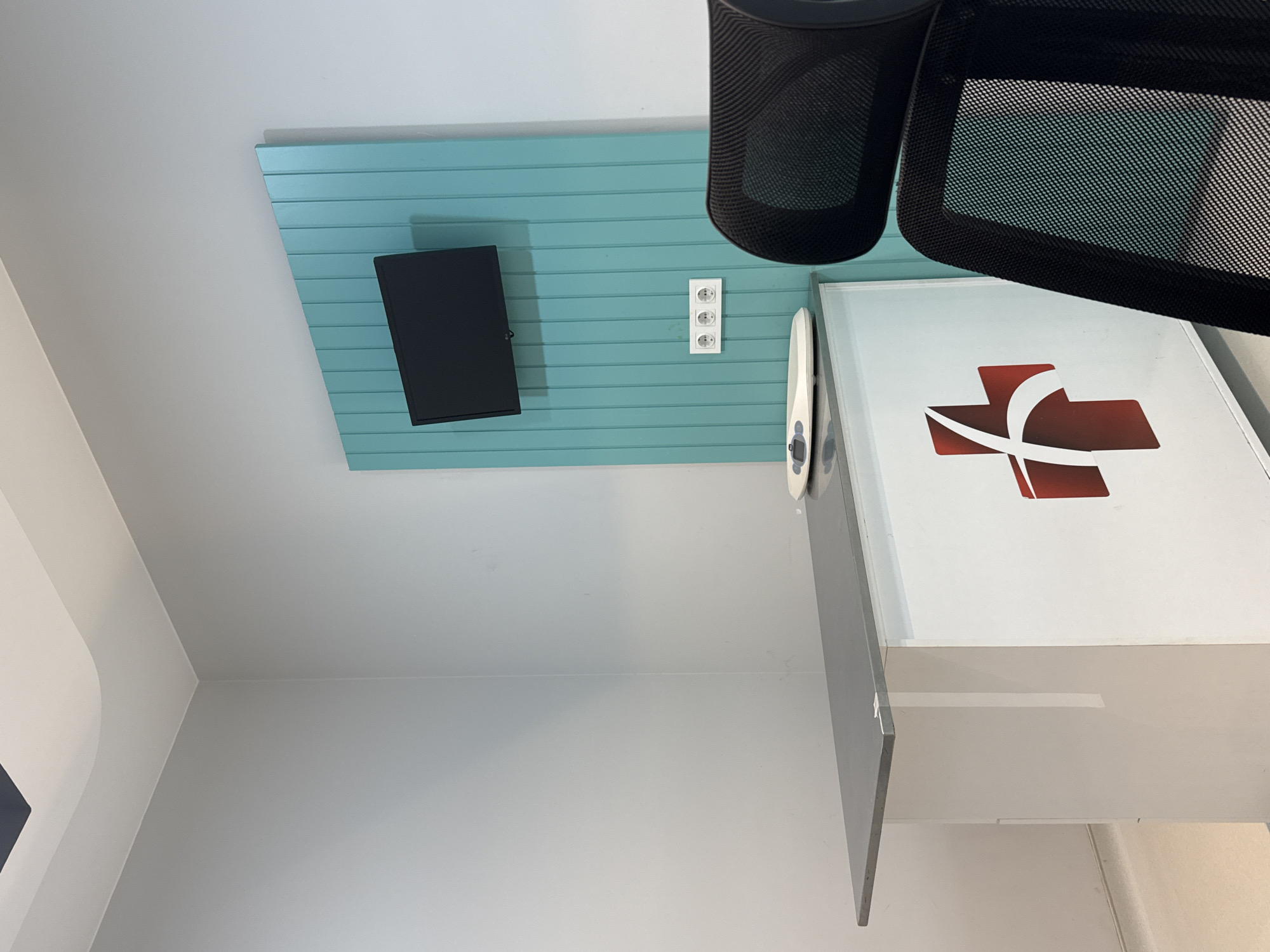 The image depicts a room with a gray wall and a light blue panel. A black flat-screen television is mounted on the panel, along with a white electrical outlet. A white cabinet with a red cross logo is next to the panel. A black office chair is visible in the foreground. The location is Podgorica, Montenegro.