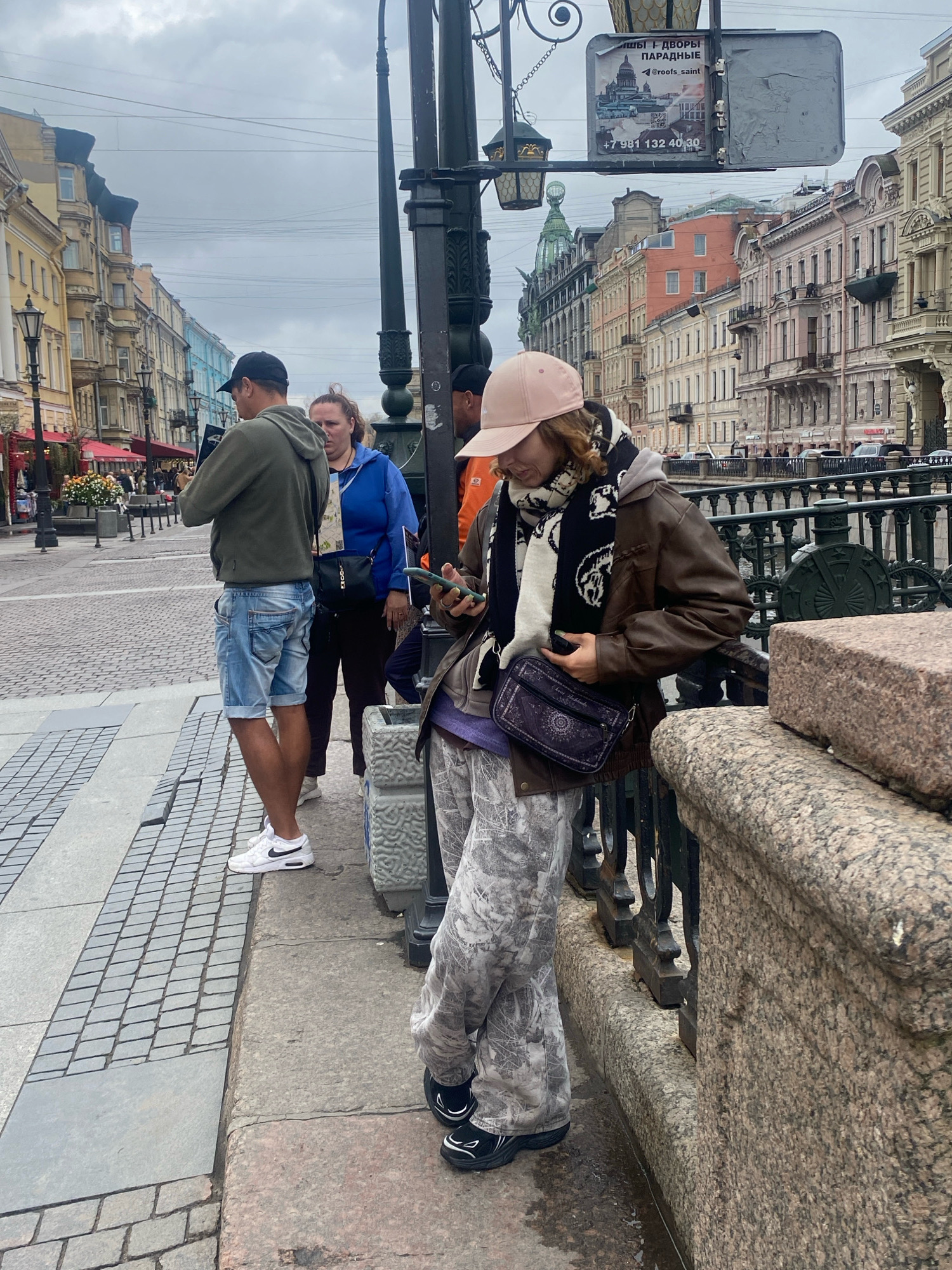 The image shows a street scene in Saint Petersburg, Russia, likely near a canal or river given the presence of a stone embankment and railing. In the foreground, a person wearing a light pink baseball cap, a brown leather jacket, a black and white scarf, and camouflage-patterned pants is looking at their mobile phone. They are leaning on the stone railing. To the left, two other individuals are visible. One, wearing a green hoodie and blue denim shorts, is also looking at a phone. Behind them, a person in a blue jacket is looking towards the left. The background features a street lined with classical European-style buildings, characterized by ornate facades and varied rooflines. The sky is overcast, suggesting a cloudy day. A street lamp post with decorative elements is prominent in the center of the image. A sign with Cyrillic text and an image of a building is attached to the lamp post. The ground is paved with cobblestones and some larger stone slabs.
