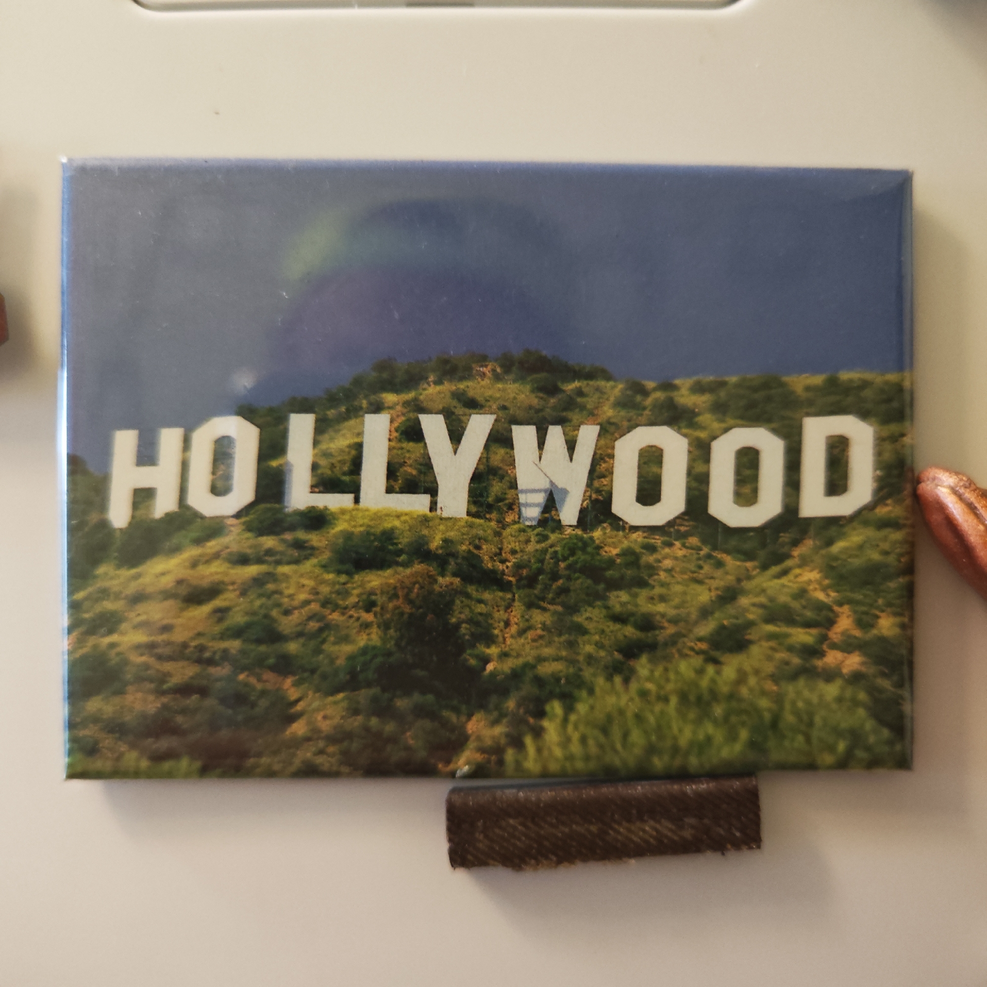The image shows a rectangular magnet depicting the Hollywood sign on a hillside under a blue and cloudy sky. The magnet is attached to a white surface. A small brown rectangular object is located beneath the magnet. A partially visible, reddish-brown object is on the right side of the magnet. The image does not contain any information relating to the city of Ketoshi, Russia.