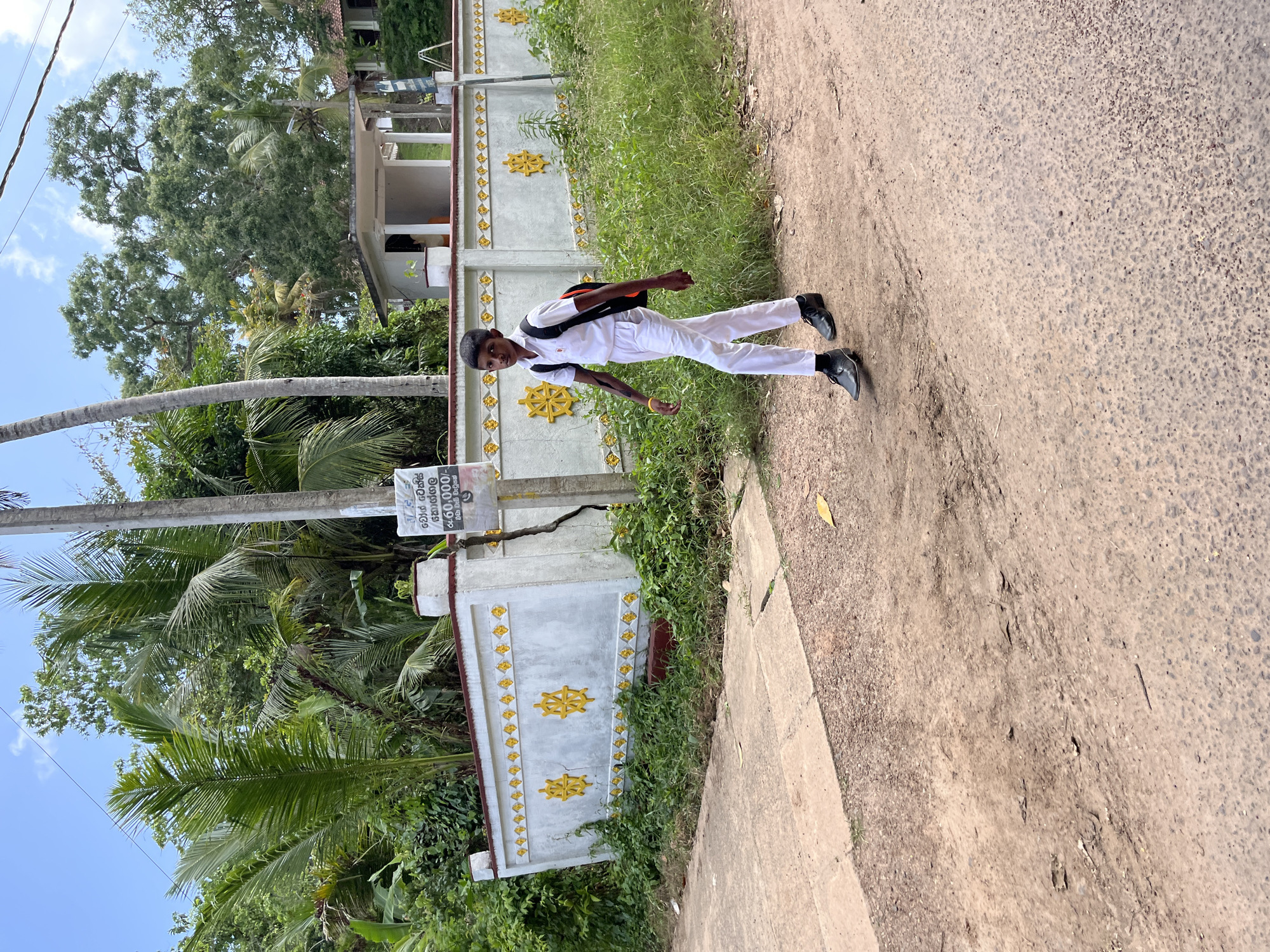 A young boy in a white school uniform walks along a dirt road in Kabalana, Sri Lanka. He carries a black and orange backpack. He is wearing black dress shoes and a yellow wristband on his left arm. The boy walks past a white wall decorated with yellow symbols. The wall is overgrown with green foliage. Palm trees and other lush vegetation are visible in the background. A small white building with a covered entrance is also visible behind the wall. The sky is blue with scattered clouds. The road is unpaved and appears dry, with patches of gravel and dirt.