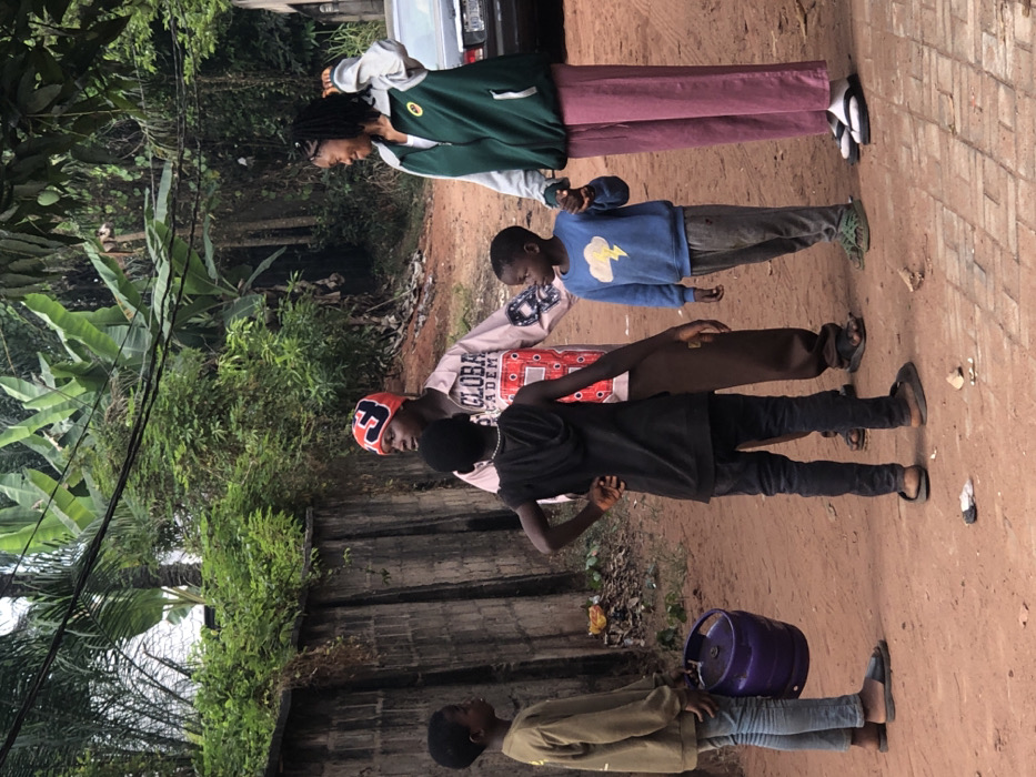Five individuals stand outdoors on a dirt path next to a building. The first person wears a green and white jacket and pink pants. The second person has on a blue sweatshirt. The third individual has on a pink long-sleeved shirt. The fourth wears black pants and a black shirt. The last individual is carrying a small keg. They appear to be standing at attention. There are trees to the left and a vehicle behind the first person.