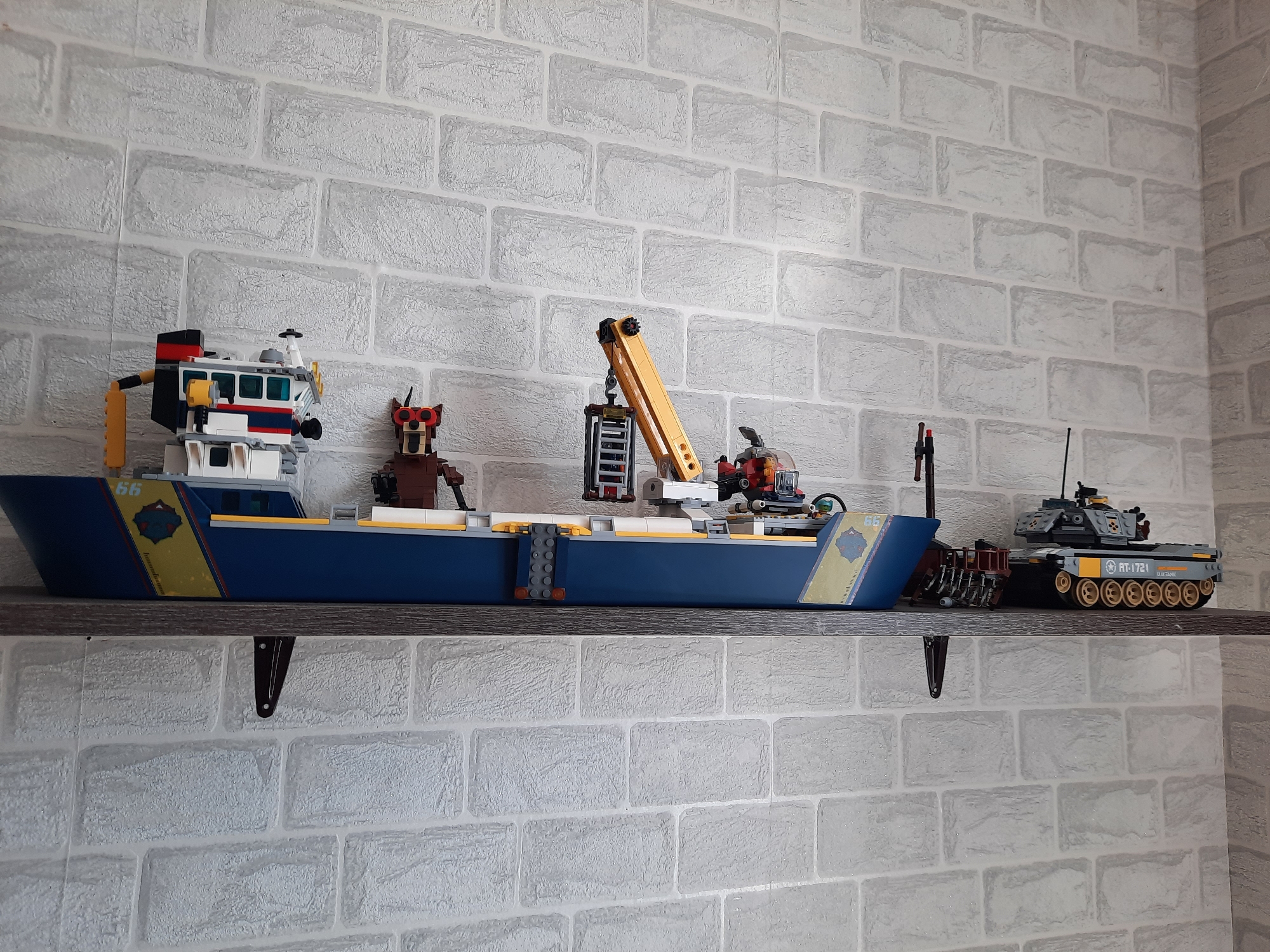 A gray shelf is mounted on a white brick wall. The shelf holds a collection of LEGO models: a blue and yellow cargo ship, a brown robot-like figure, and a gray tank labeled "RT-1721." The location is not discernable from the image.