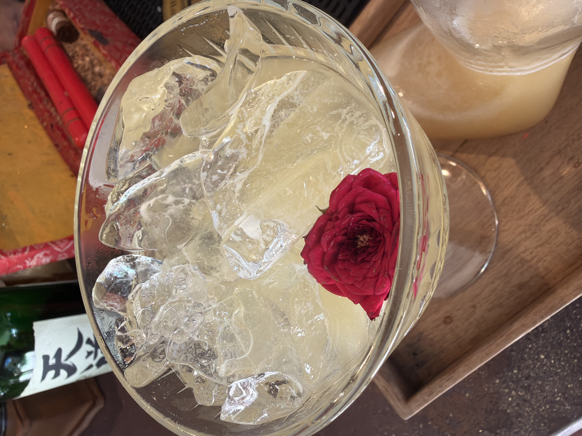 A close-up, overhead shot captures a cocktail filled with ice and a single red rosebud. The drink is served in a clear, stemmed glass set on a wooden tray. In the background, a green bottle with Asian characters and a blurred red box containing red cylindrical objects can be seen. The scene appears to be indoors, possibly a bar or restaurant, located in Baan Thong Krut, Thailand.
